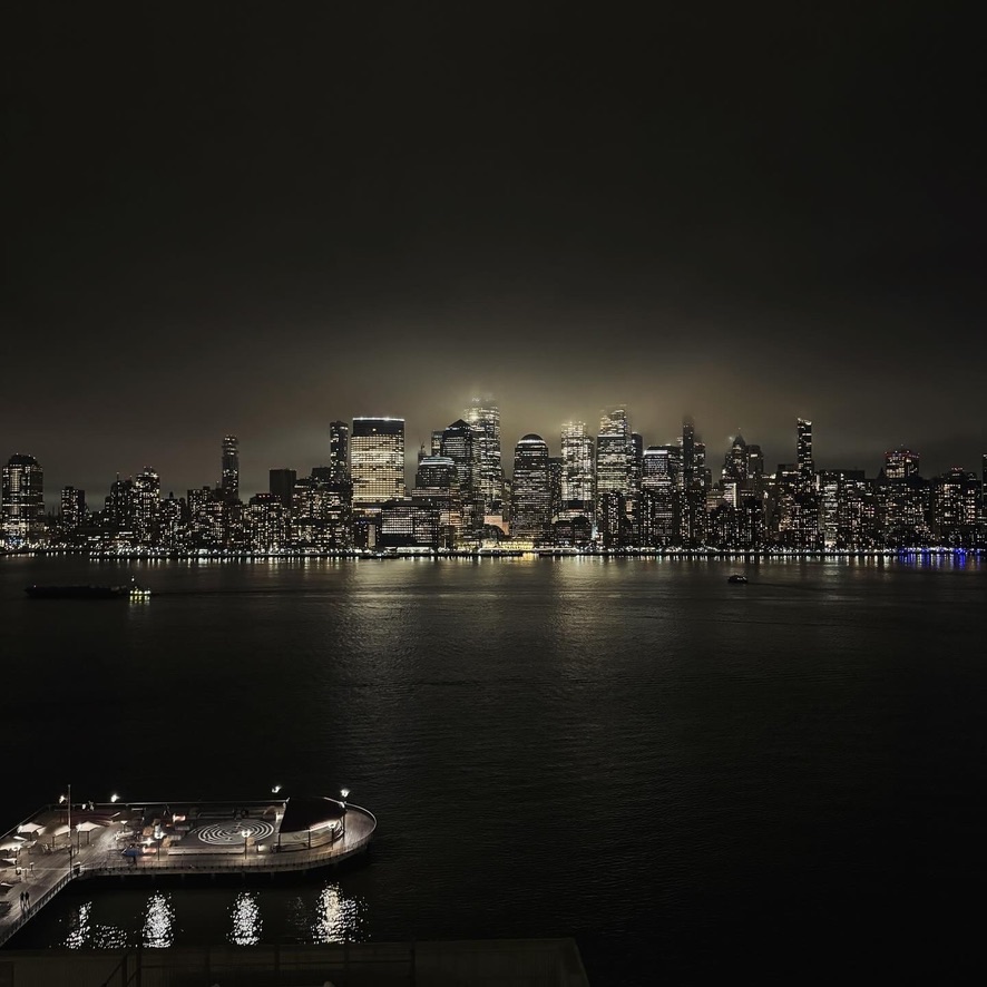 Lower Manhattan skyline at night, lit towers reflected in the dark river, a circular pier in the foreground