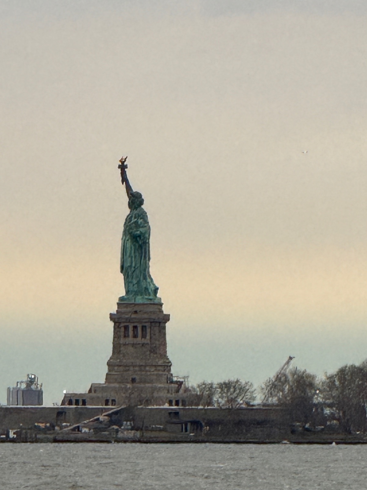 The Statue of Liberty green against a pale gold evening sky, seen across the water from the Battery Park City esplanade