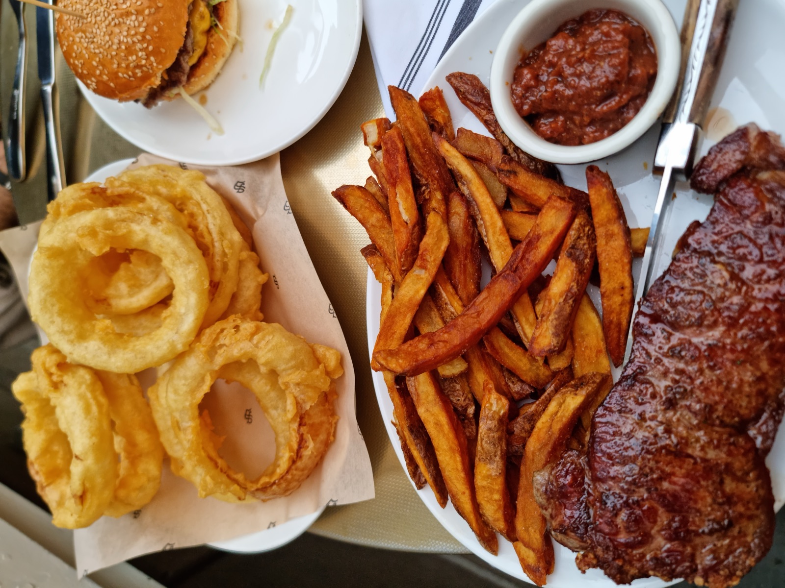 A grilled steak with thick hand-cut fries, a stack of onion rings on a separate plate and a pot of dark sauce