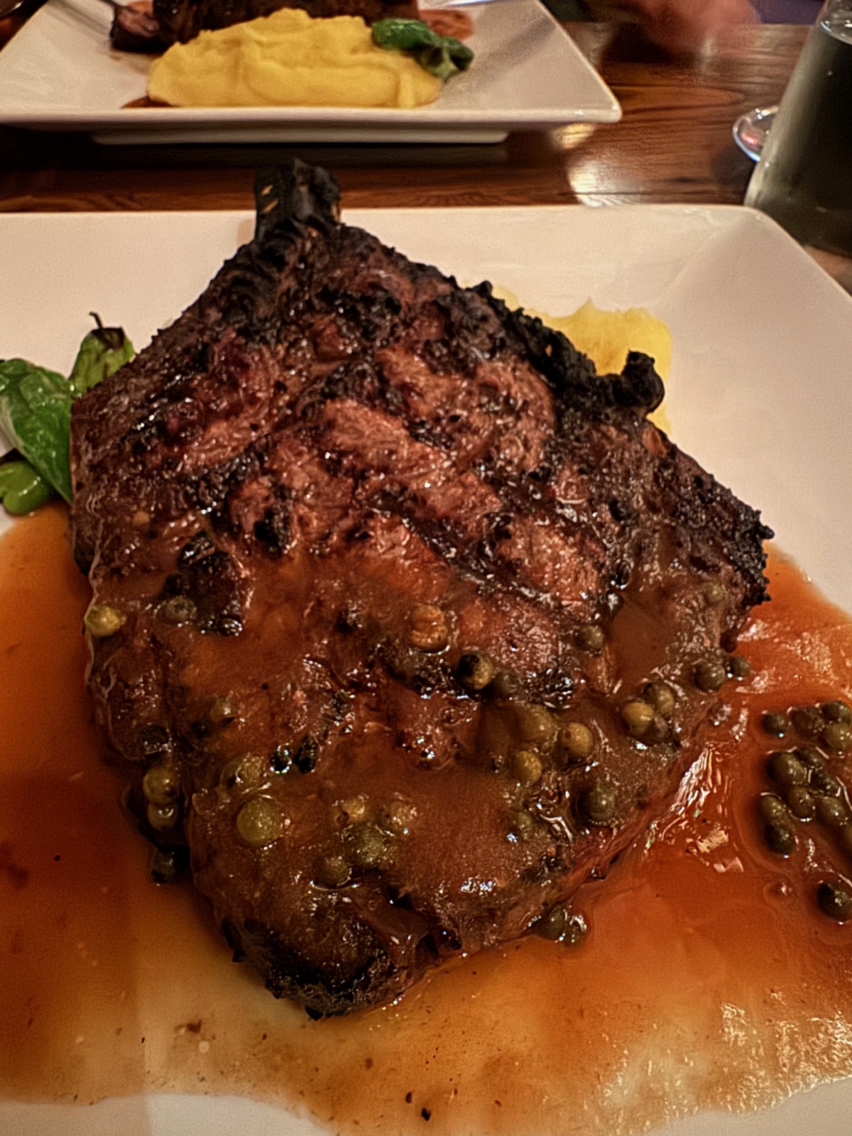A thick grilled rib of beef on a white plate in a dark green peppercorn sauce, with mashed potato on a plate behind