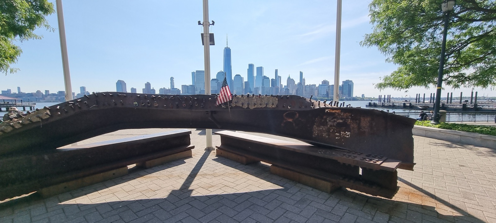 A twisted rust-brown steel beam mounted on a low plinth on a paved waterfront, a small United States flag behind it and the Lower Manhattan skyline across the water