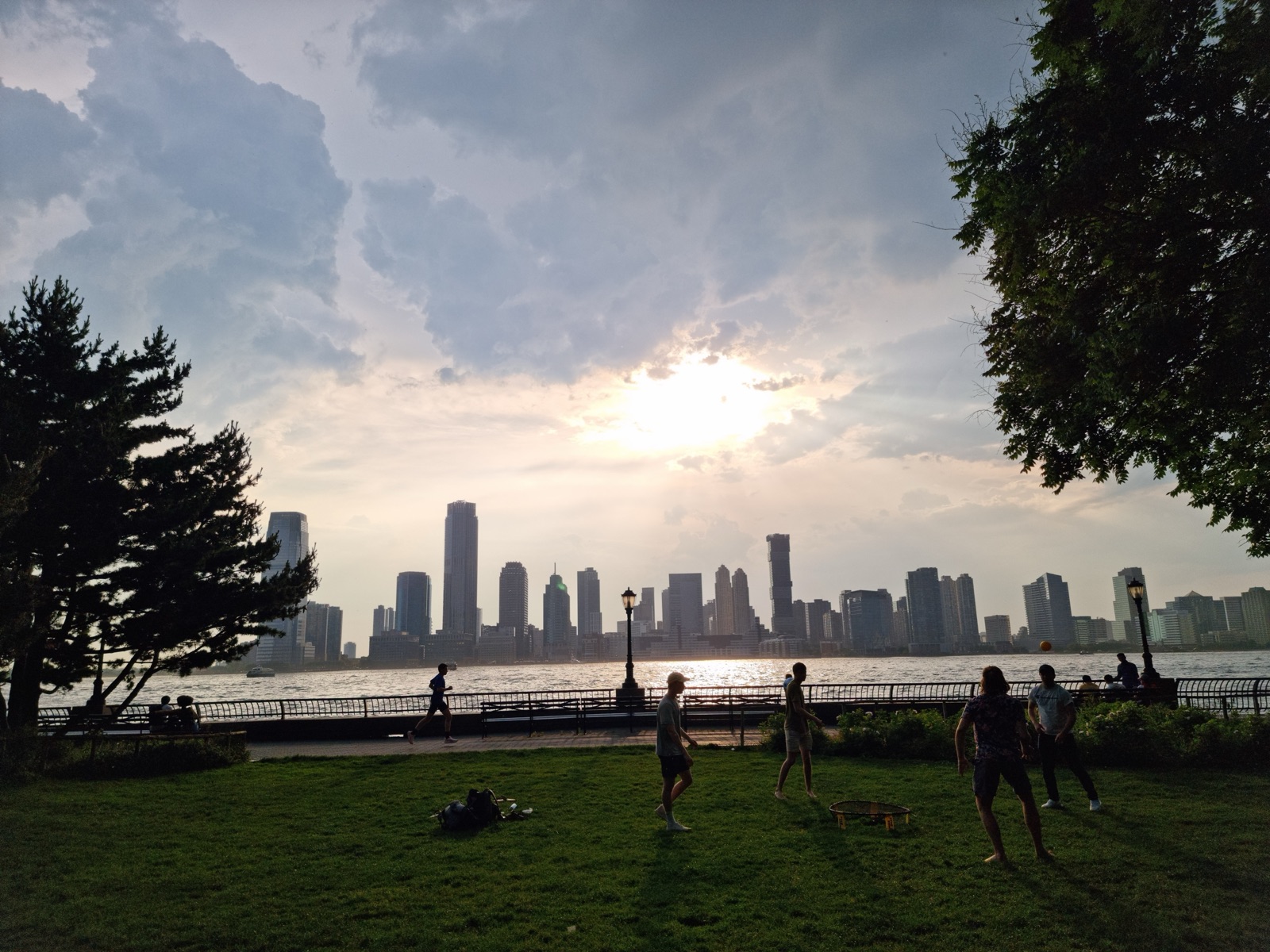 The low afternoon sun behind cloud over the Jersey City skyline seen across the Hudson from a grassy park, with trees framing the view and people playing on the lawn