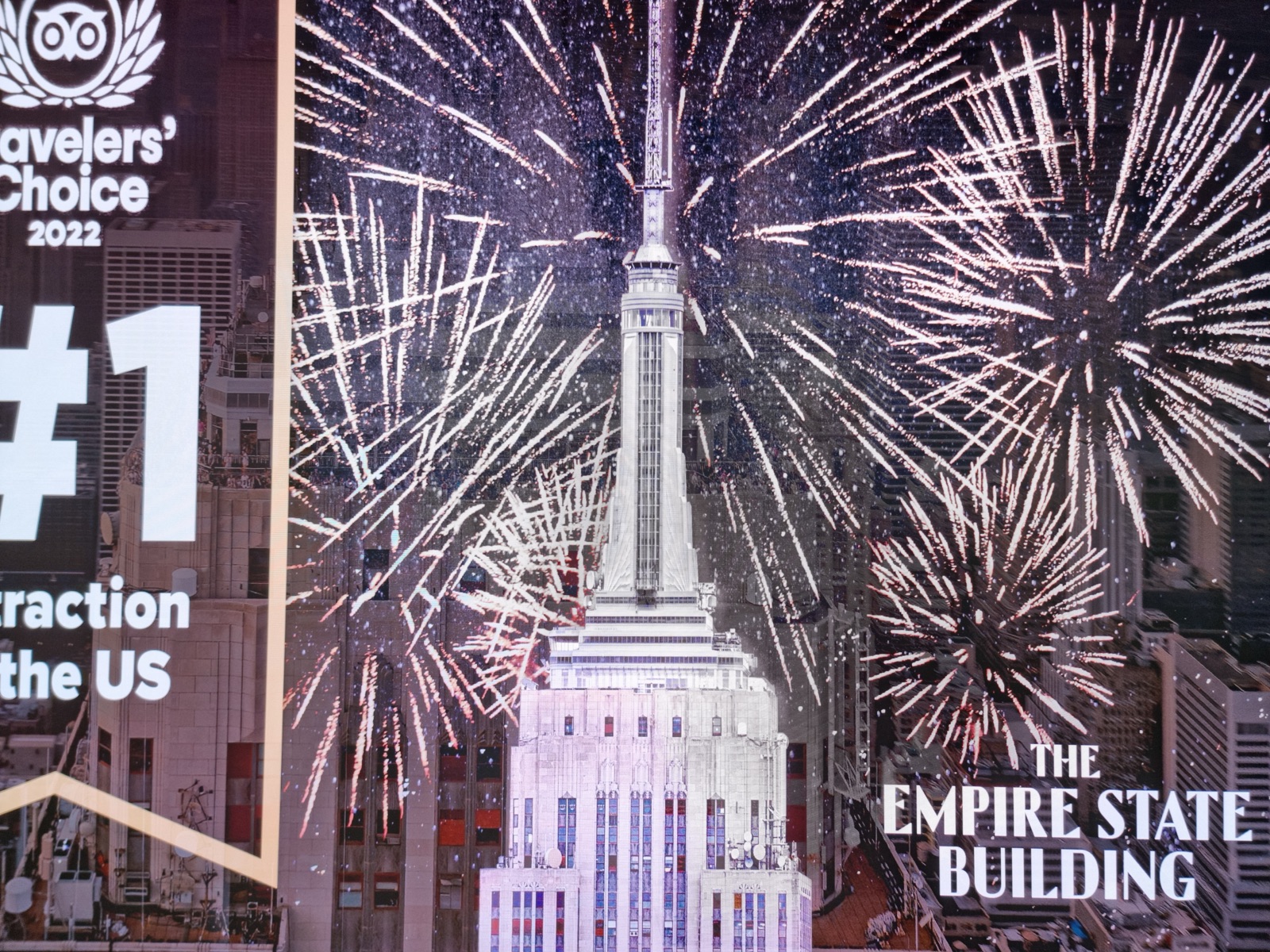 An illuminated advertising panel reading THE EMPIRE STATE BUILDING beside a photograph of the tower lit up with fireworks bursting around it