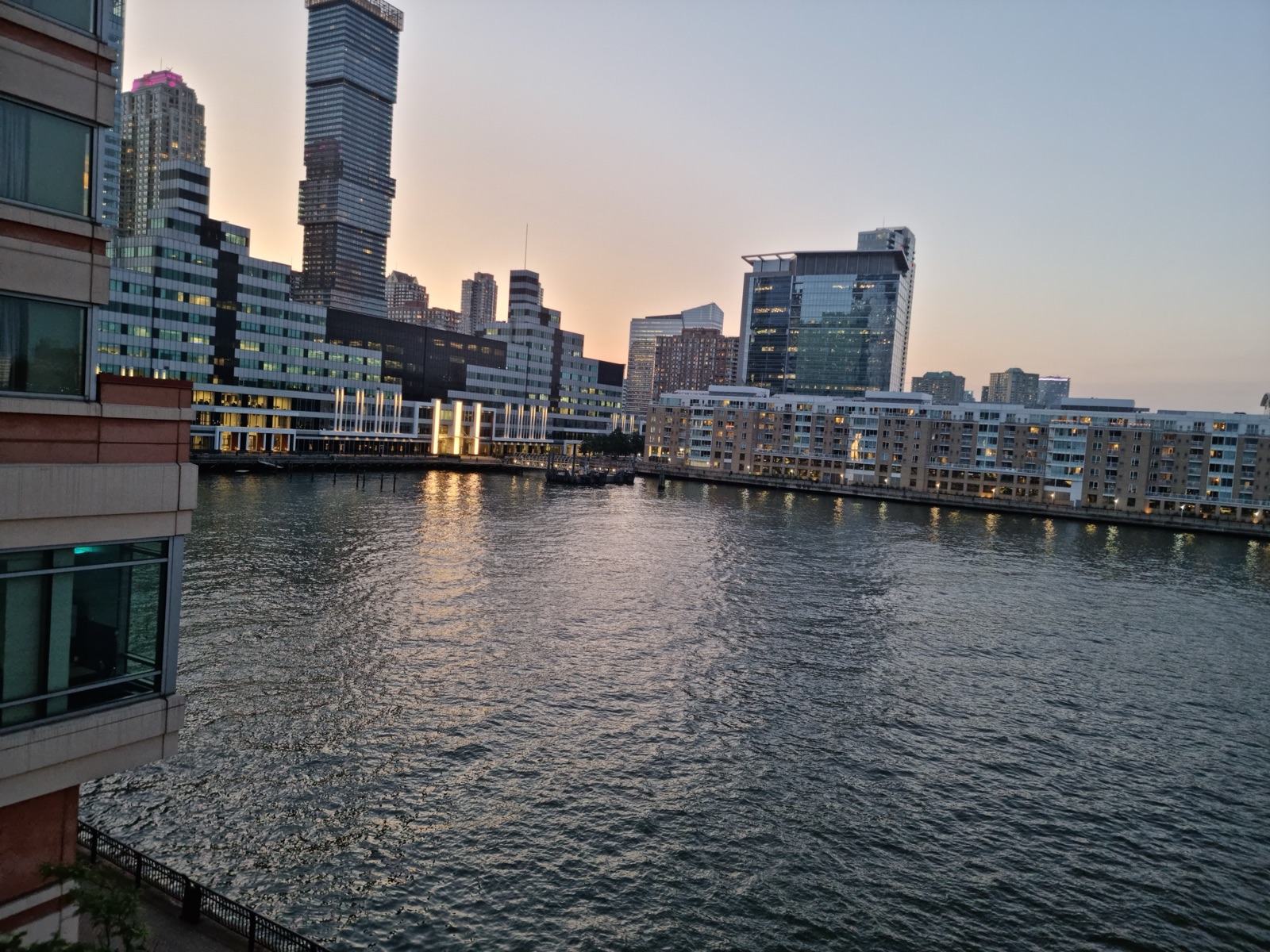 The Jersey City waterfront buildings and the river at dusk with the Manhattan skyline low and lit on the far side