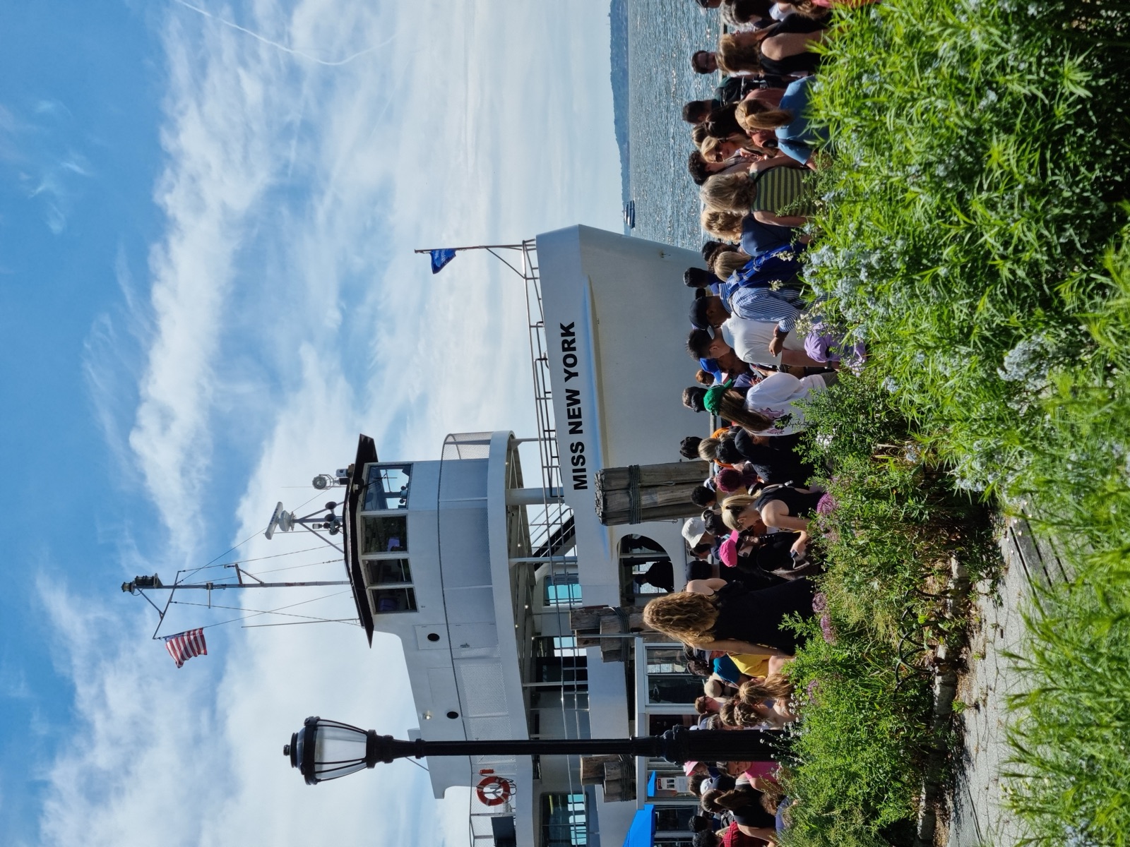 A white harbor ferry named MISS NEW YORK moored beyond a crowd of people waiting on a planted waterfront path