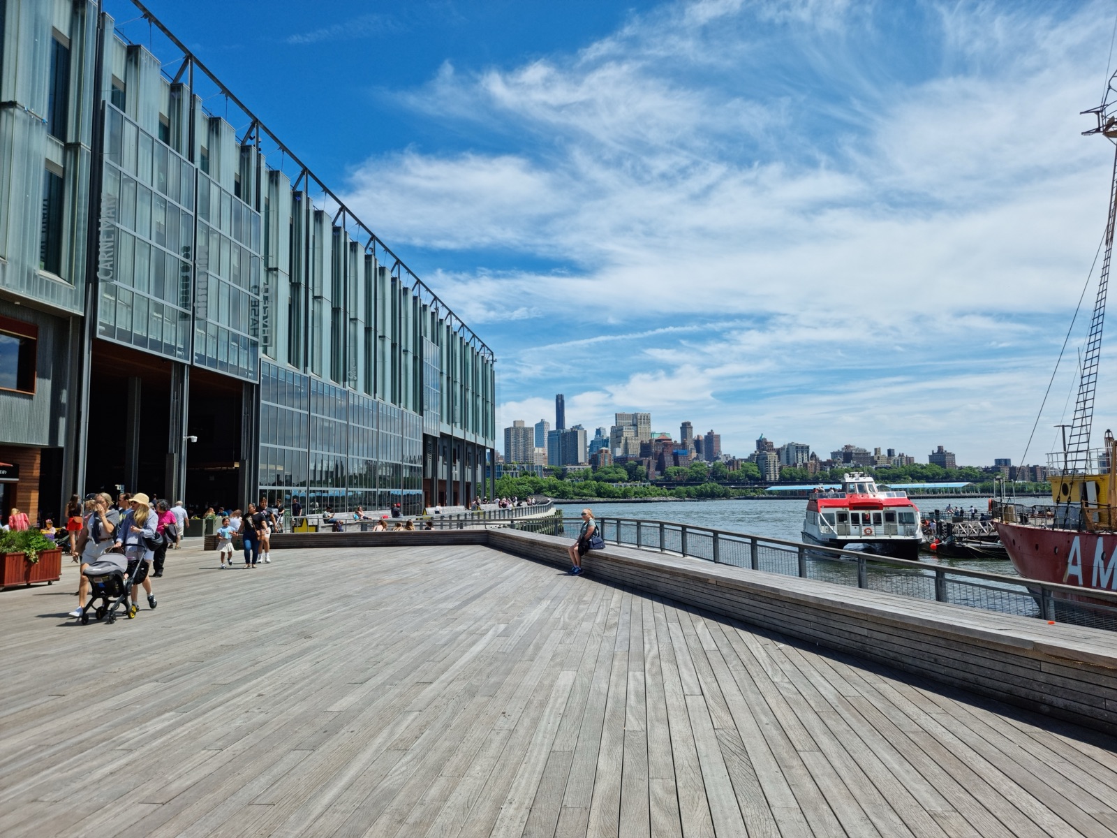 A wide timber pier at South Street Seaport with people strolling, a glass building on the left and a red-hulled boat and the Brooklyn Bridge beyond