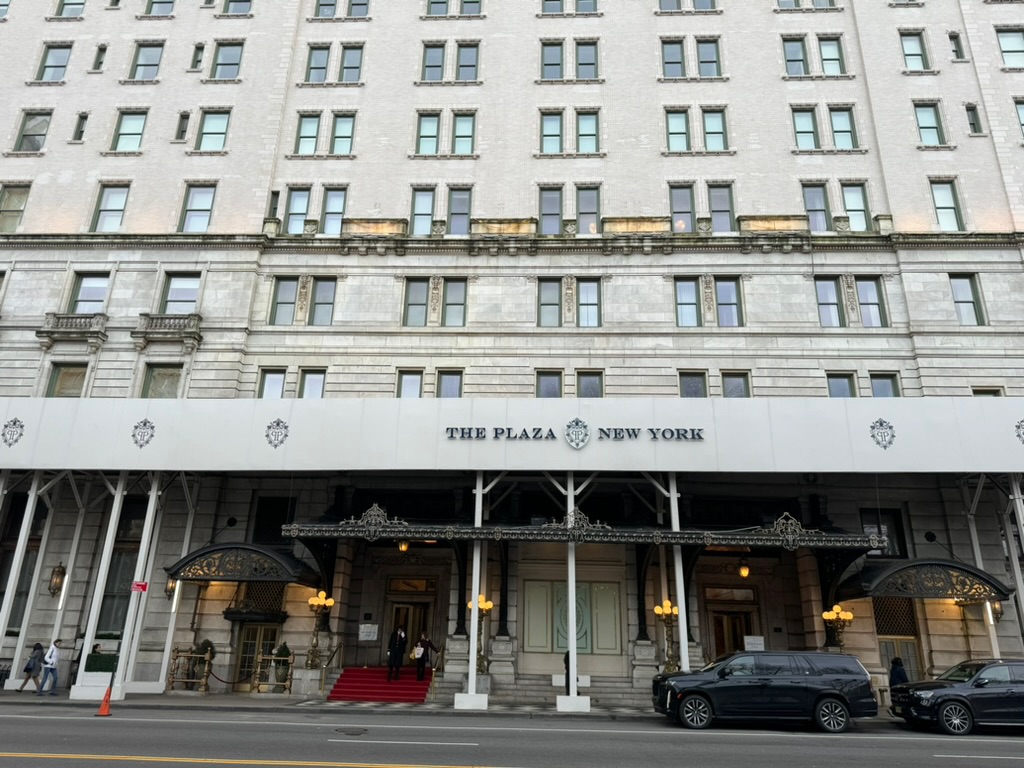 The limestone facade and awning of the Plaza Hotel, its name lettered above a red-carpeted entrance