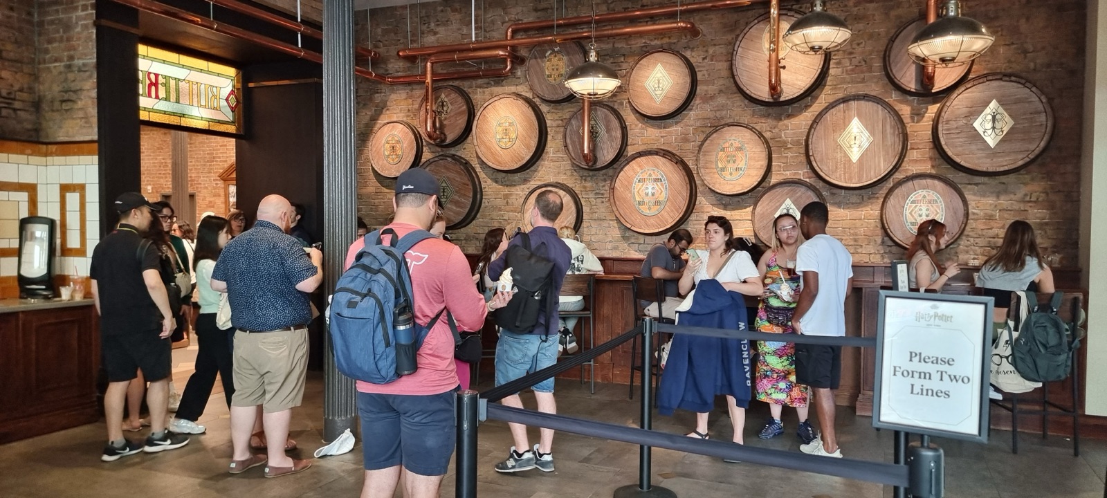 A line of people waiting between rope barriers inside Harry Potter New York, below a brick wall hung with the ends of wooden barrels stamped with butterbeer crests, copper pipes and brass lamps, and a sign reading Please Form Two Lines