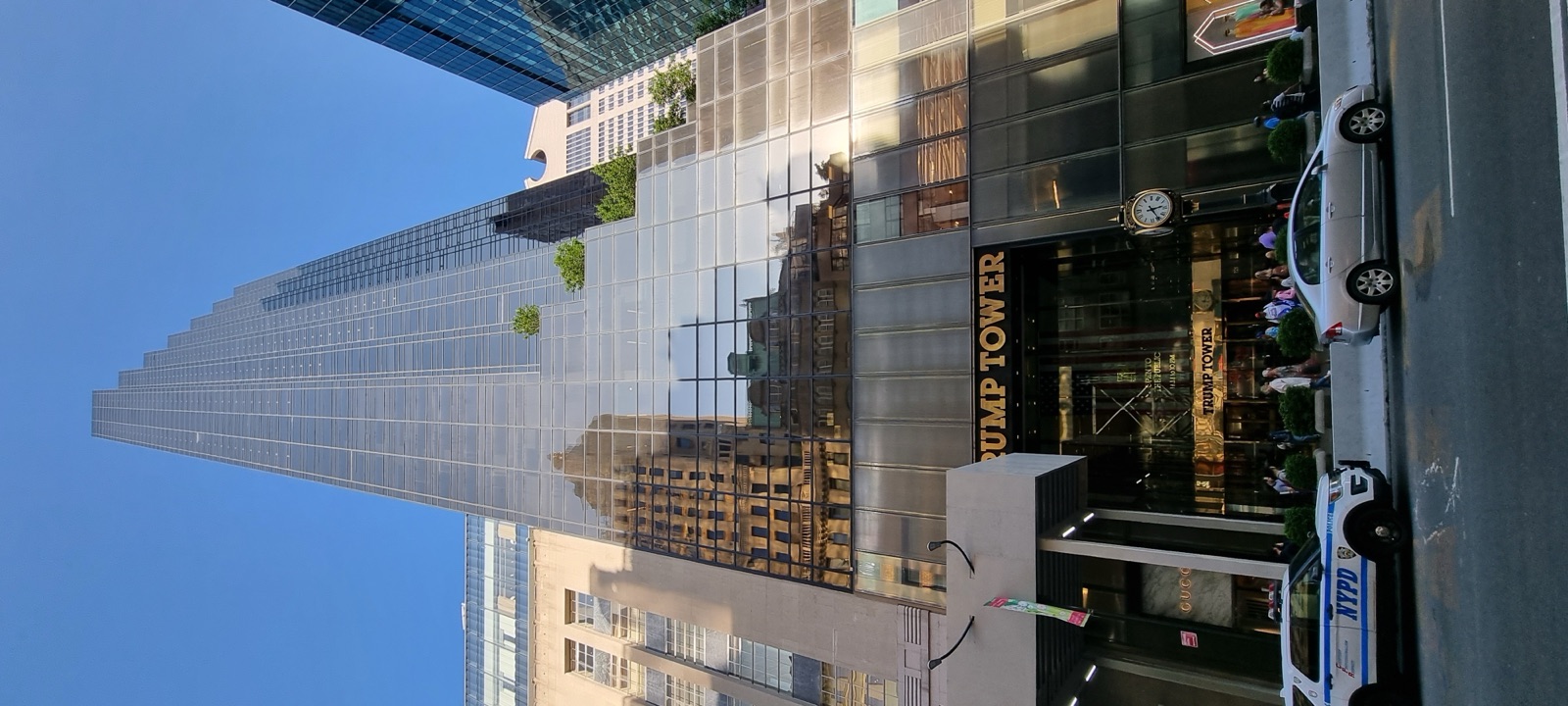 A tall dark glass tower on Fifth Avenue seen from the street below, with a police car parked at the entrance