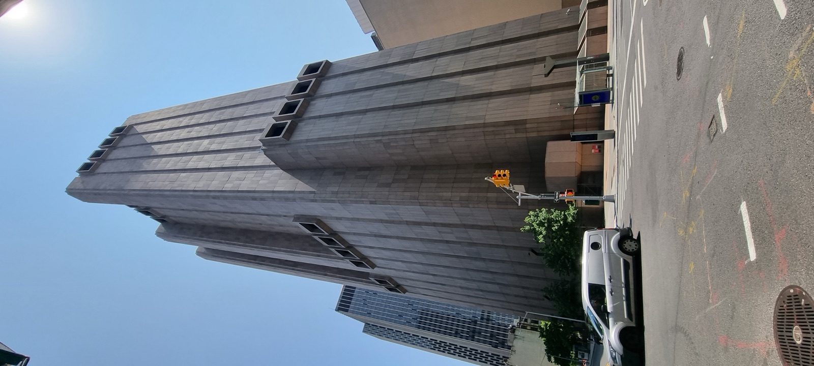 A tall windowless concrete tower with narrow ventilation slots rising sheer from the street against a blue sky, a glass building beside it