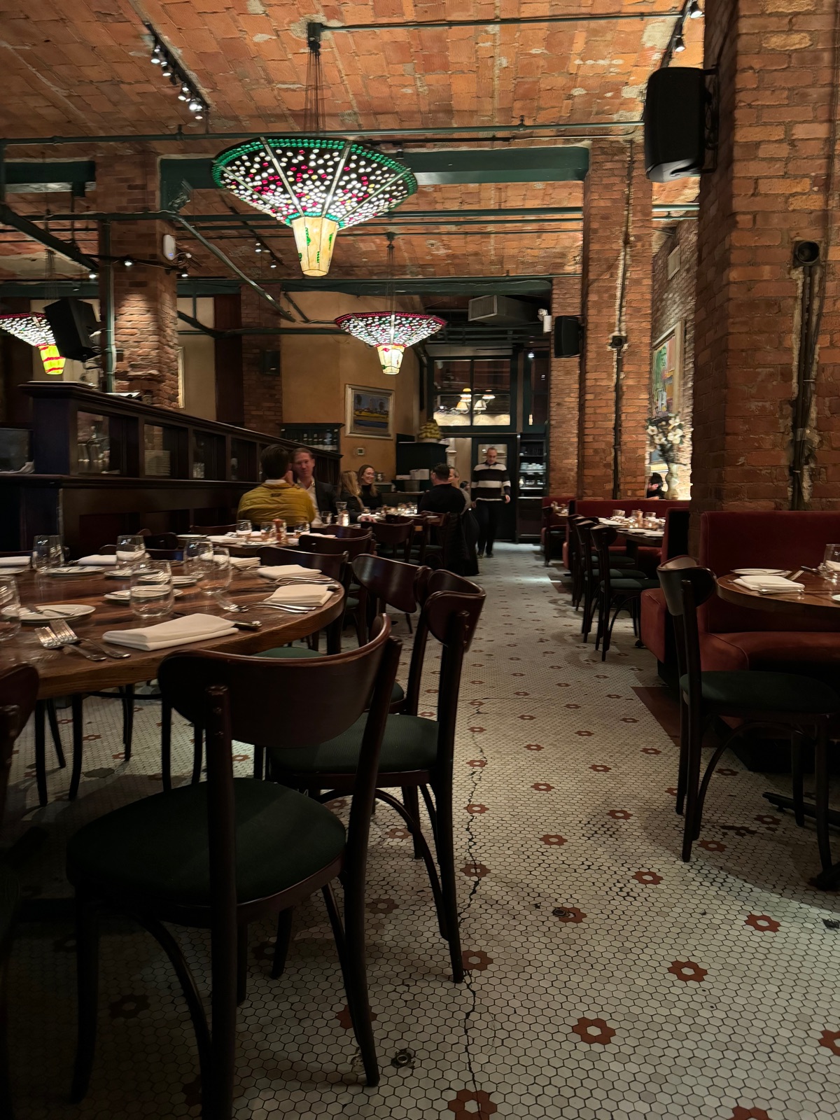 A dining room with exposed brick vaults, green painted steel beams, stained glass pendant shades and a mosaic tile floor, tables set with white napkins