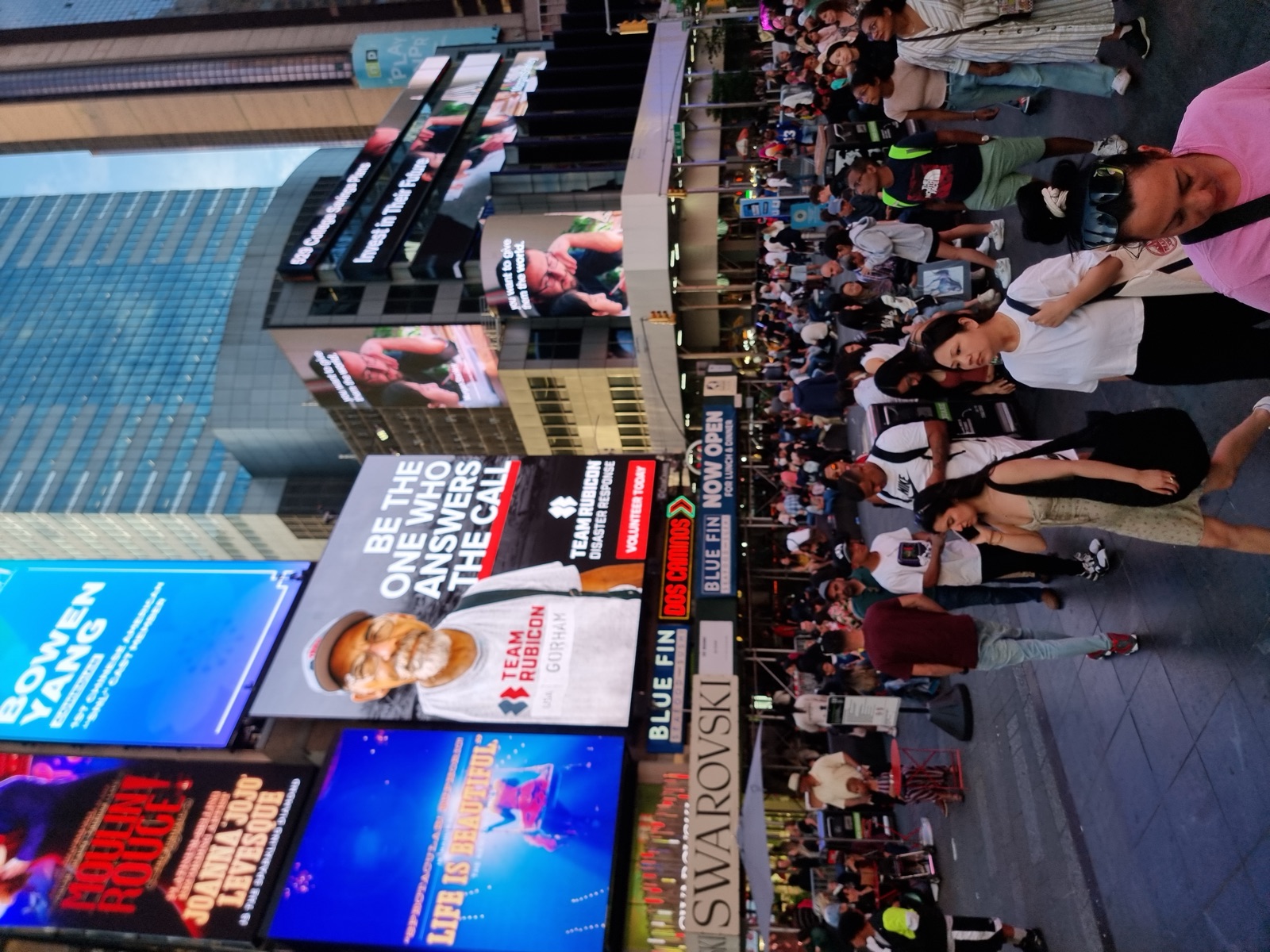 A dense crowd of people crossing Times Square in the evening beneath lit advertising screens on every building