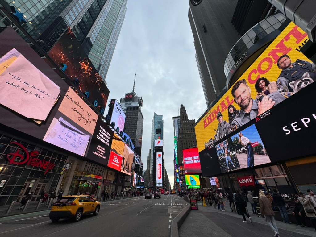 Times Square at dusk, glowing billboards stacked on every building, a yellow cab and crowds beneath a wide grey sky