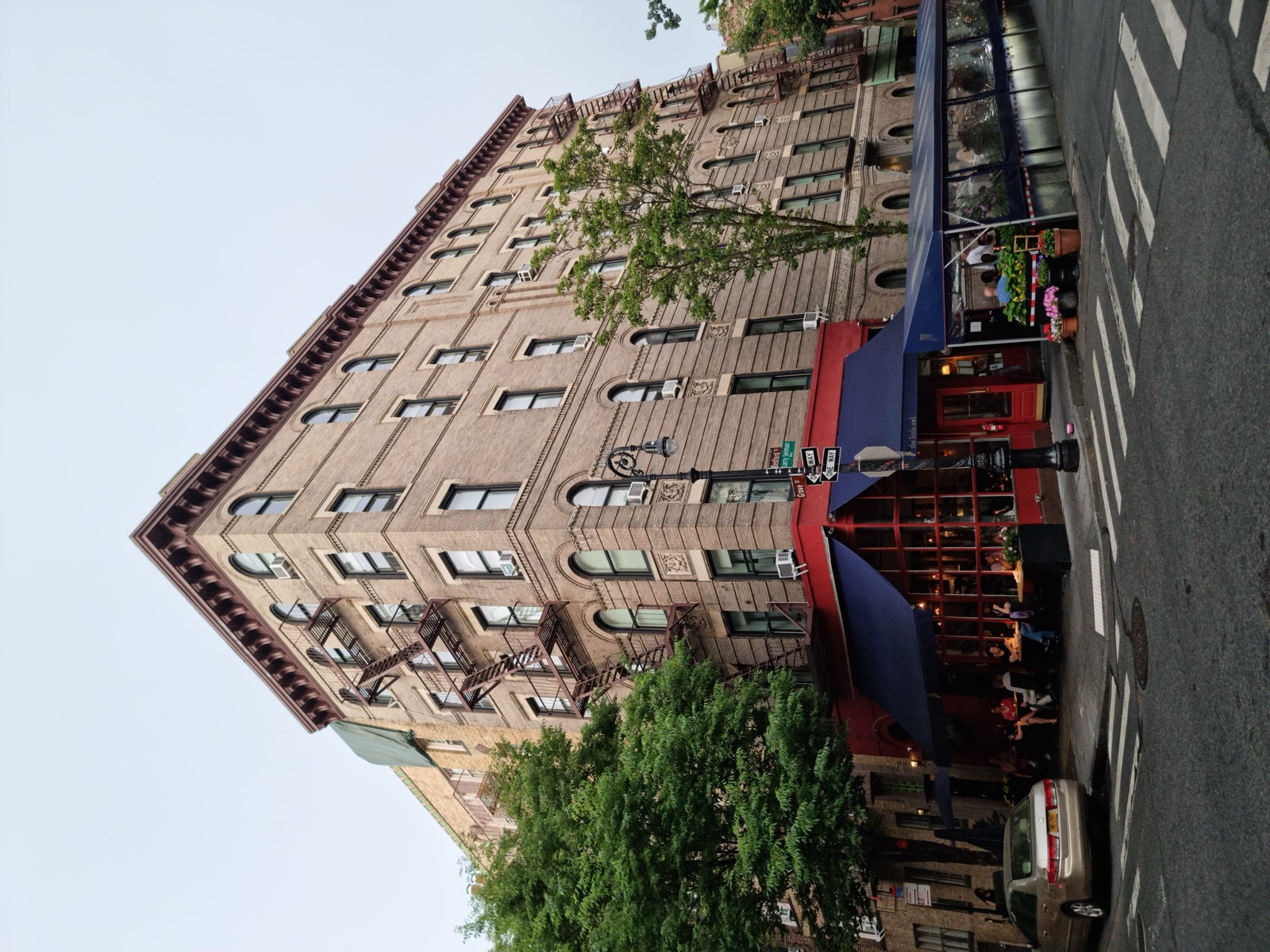 The tan brick corner building at Bedford and Grove Streets in Greenwich Village, fire escapes zigzagging down its front, with The Little Owl restaurant in red under a blue awning on the ground floor and leafy green trees on both streets