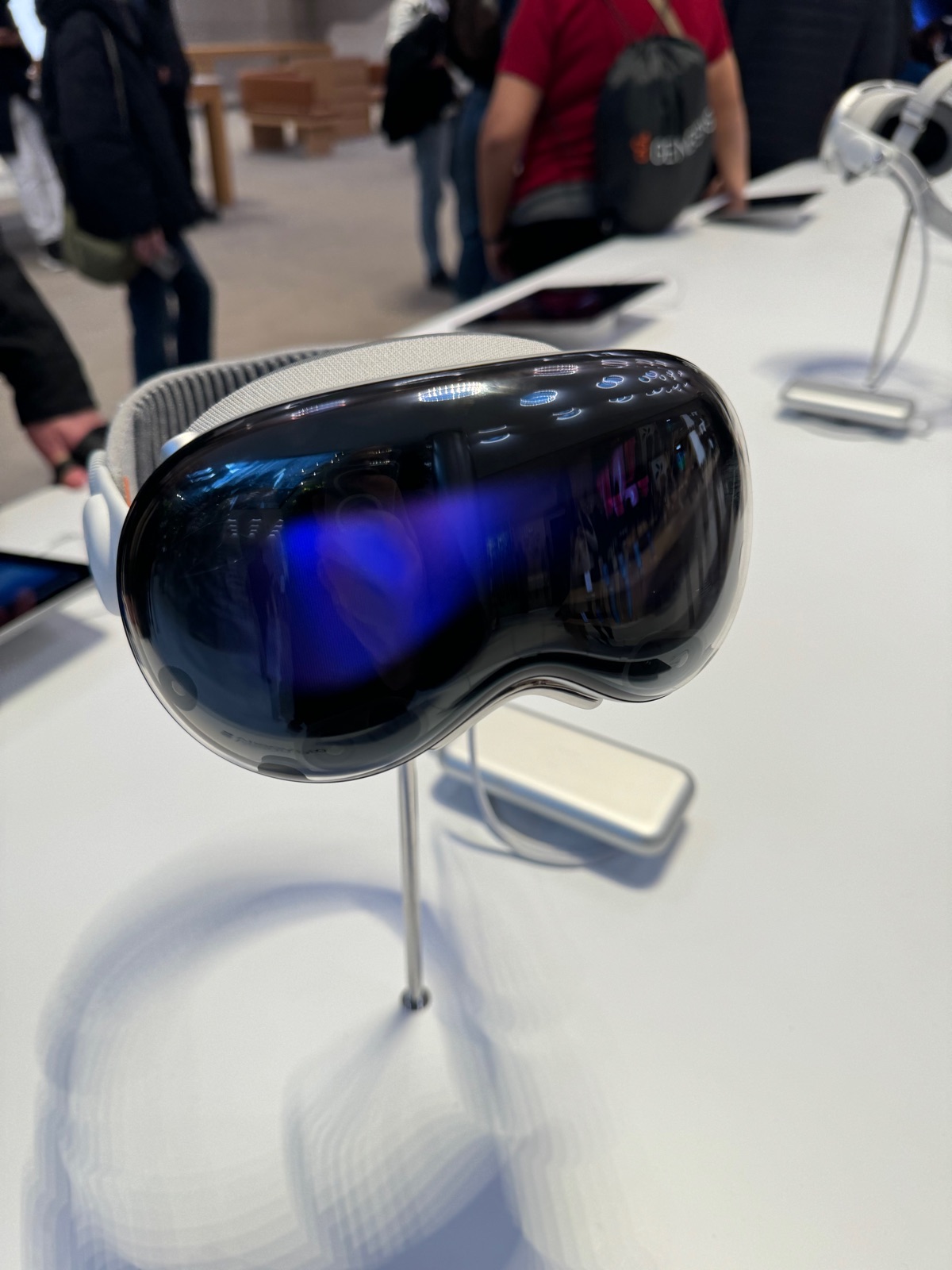An Apple Vision Pro headset on a display stand on a white table in a busy store