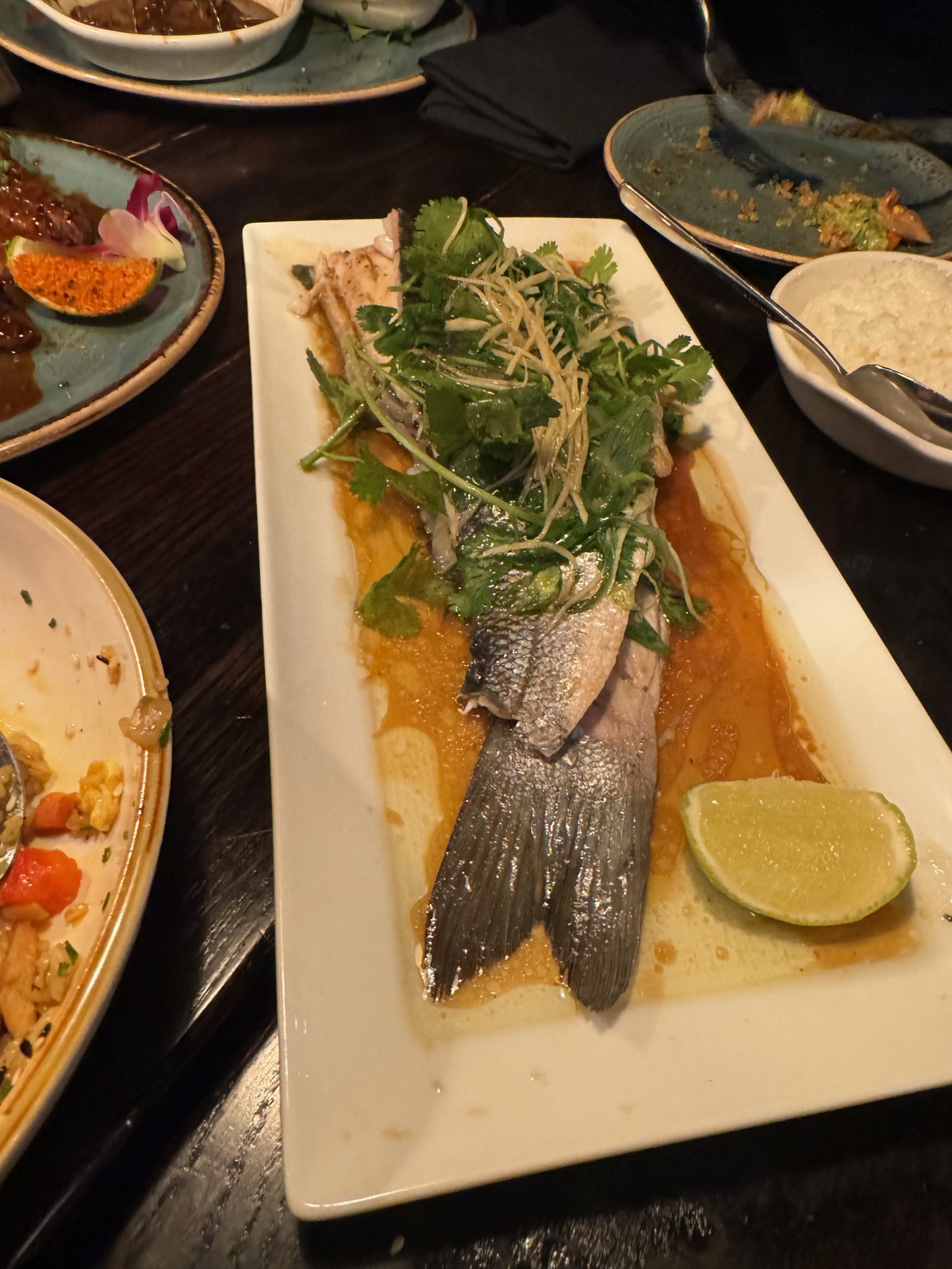 A whole fish on a long white plate in an orange broth, topped with cilantro and shredded scallion with a wedge of lime