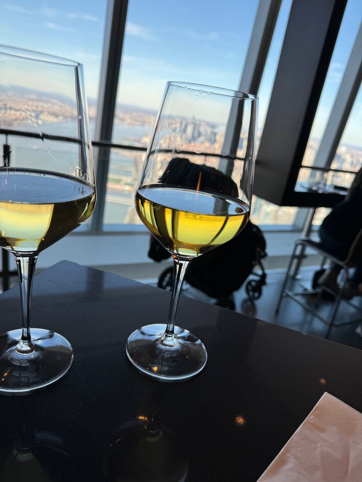 Two glasses of white wine on a dark table against the sloping window of an observation deck, the city and the harbor far below outside