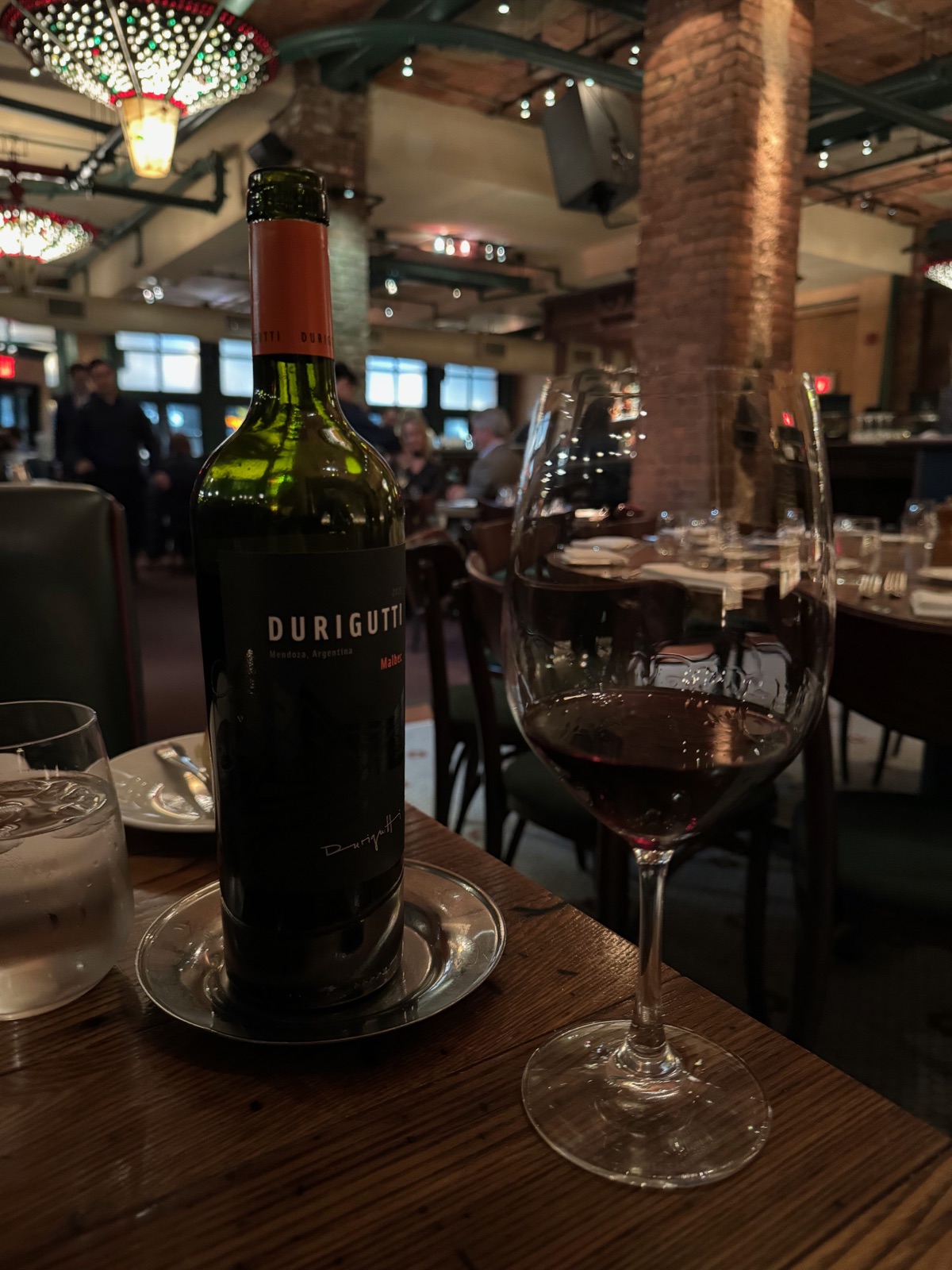 A bottle of Durigutti Malbec and a poured glass of red wine on a wooden table in a warmly lit dining room