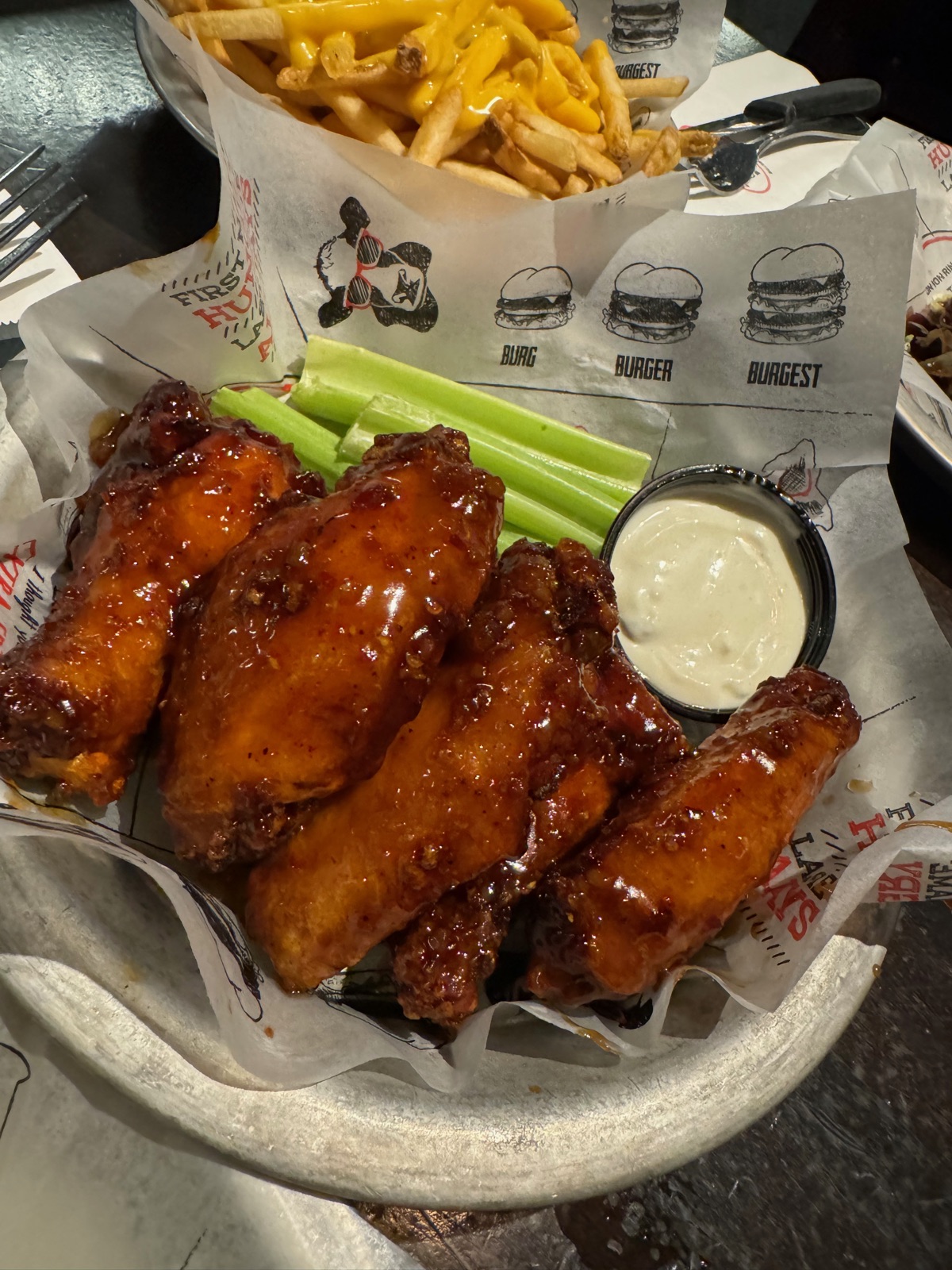 A basket of glazed chicken wings with celery sticks and a pot of blue cheese dip, beside cheese fries in paper printed with burger sizes