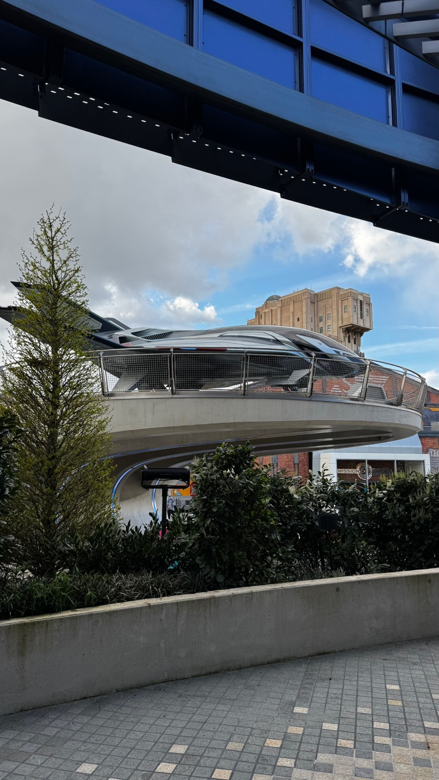 Ένα γκρίζο quinjet παρκαρισμένο σε μια καμπύλη τσιμεντένια ράμπα στο Avengers Campus, με τον μπεζ πύργο του Hollywood Tower Hotel να υψώνεται πίσω