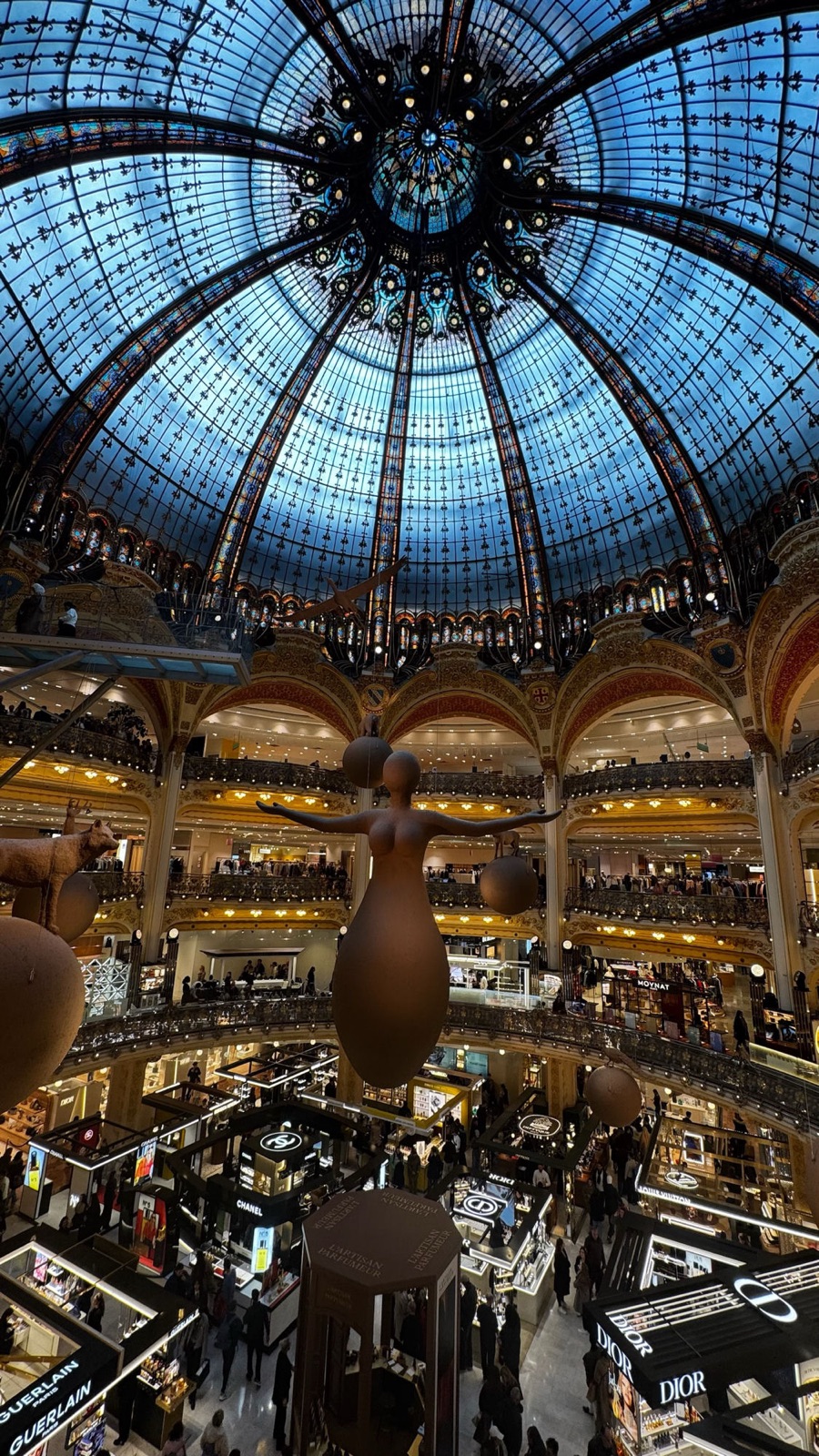 Ο μπλε και χρυσός βιτρό τρούλος των Galeries Lafayette από κάτω, με μια γλυπτή μορφή με απλωμένα χέρια αιωρούμενη πάνω από τα μπαλκόνια της αίθουσας αρωμάτων