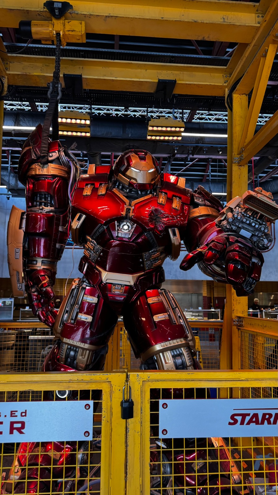 Η τεράστια κόκκινη και χρυσή πανοπλία Hulkbuster να στέκεται σε μια κίτρινη βιομηχανική γερανογέφυρα πάνω από ένα χώρο εστίασης στο Avengers Campus