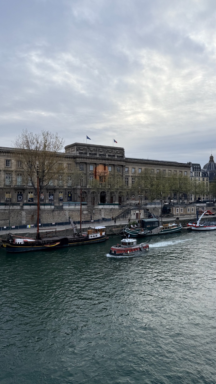 Η μακριά νεοκλασική πρόσοψη της Monnaie de Paris προς το ποτάμι το σούρουπο, με μαούνες αγκυροβολημένες κατά μήκος της προκυμαίας και ένα μικρό κόκκινο ρυμουλκό να περνά στον Σηκουάνα