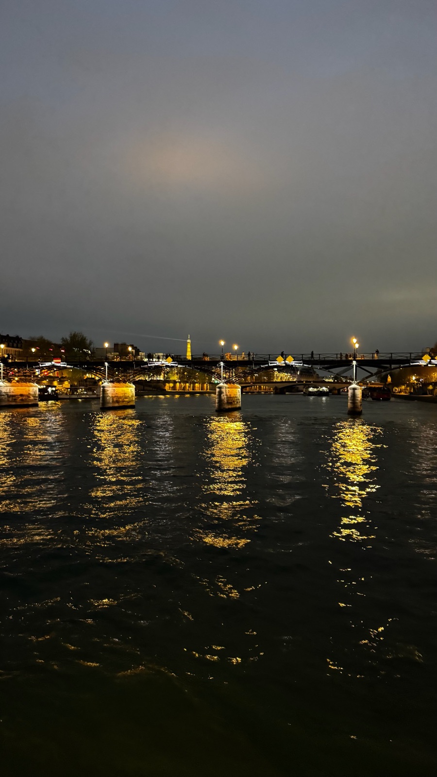 Η πεζογέφυρα Pont des Arts τη νύχτα με φωτιζόμενες λάμπες να αντανακλώνται στον σκοτεινό Σηκουάνα, με τον φωτισμένο Πύργο του Άιφελ μικροσκοπικό στον ορίζοντα