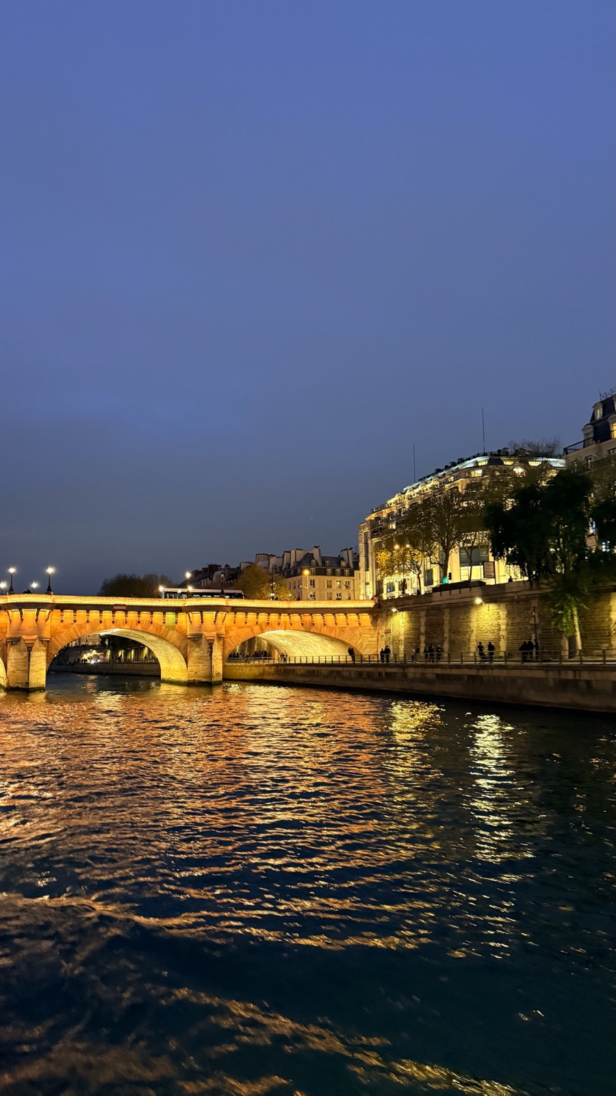 Οι φωτισμένες πέτρινες αψίδες της Pont Neuf να αντανακλούν χρυσές στον Σηκουάνα το σούρουπο, με φωτισμένα κτίρια κατά μήκος της δεξιάς όχθης