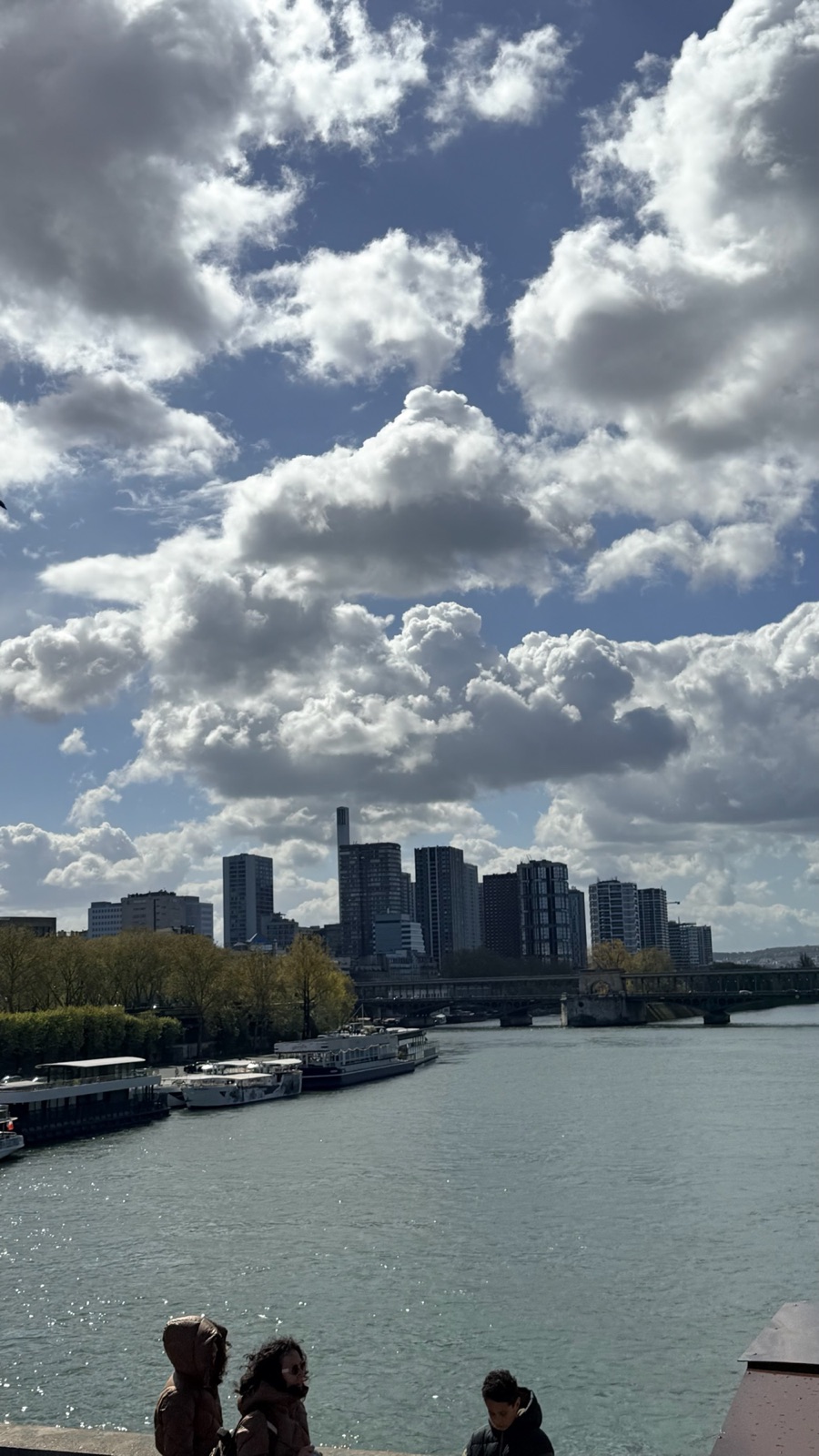 Ψηλοί πύργοι της συνοικίας Beaugrenelle στον Σηκουάνα κάτω από μεγάλα λευκά σύννεφα, με πλοιάρια κρουαζιέρας αγκυροβολημένα κατά μήκος της αριστερής όχθης