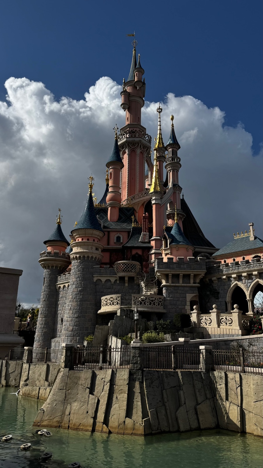 Το ροζ Κάστρο της Ωραίας Κοιμωμένης της Disneyland Paris με μπλε πυργίσκους και χρυσές αιχμές πάνω σε σκούρο μπλε ουρανό και πανύψηλα λευκά σύννεφα