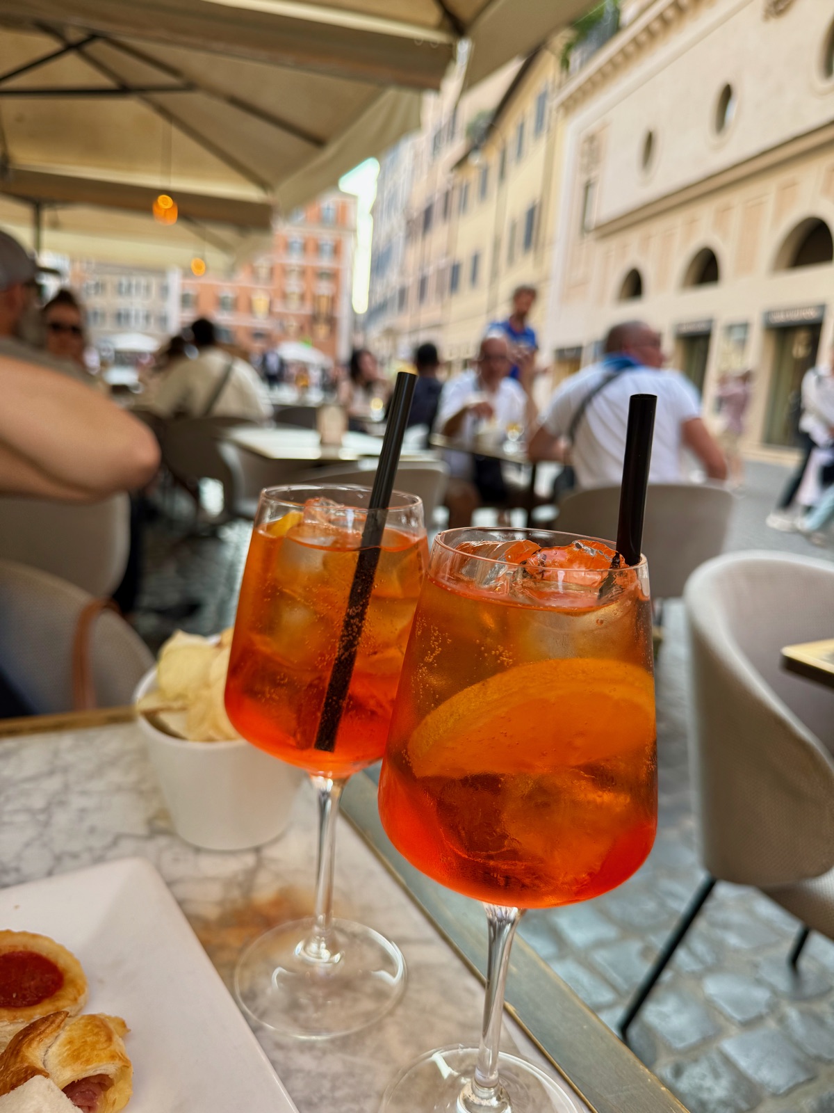 Δύο πορτοκαλί spritz με μαύρα καλαμάκια σε ένα μαρμάρινο τραπέζι καφέ κάτω από τέντα, ένα ποτήρι με γλυκό και ένα πιάτο δίπλα