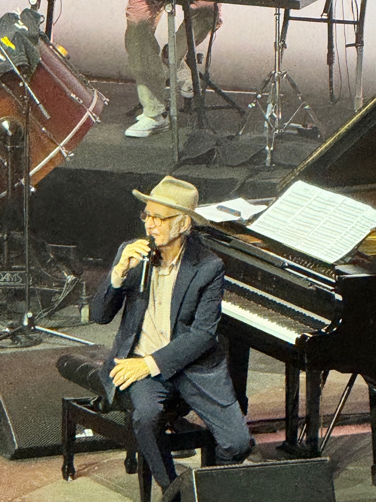 Ο Ludovico Einaudi καθισμένος στο πλάι σε ένα πιάνο με ουρά με καπέλο και σκούρο σακάκι, να μιλάει σε ένα μικρόφωνο χειρός ανάμεσα στα κομμάτια