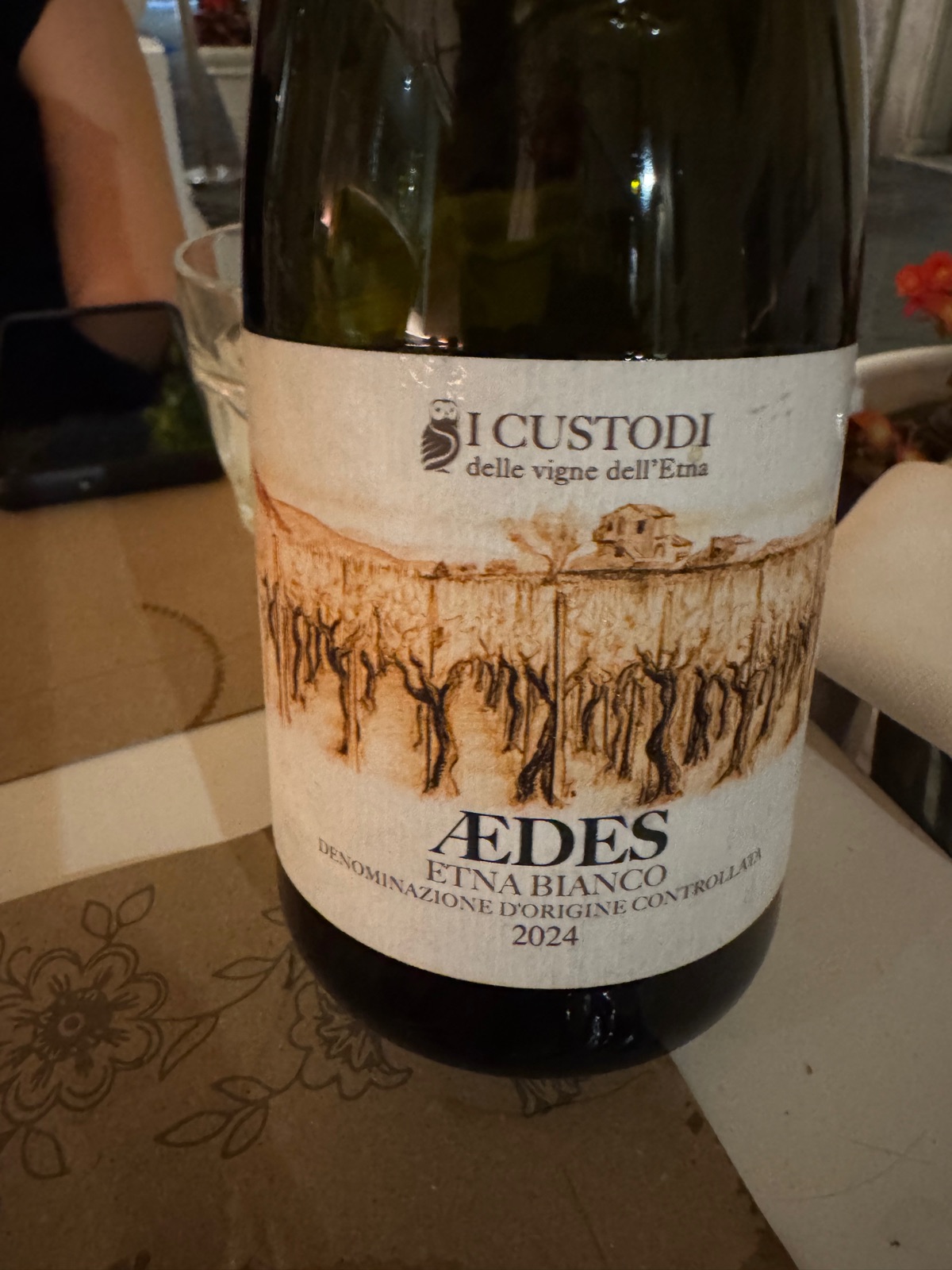 Ένα μπουκάλι Aedes Etna Bianco 2024 από τους I Custodi delle vigne dell'Etna πάνω σε ένα τραπέζι, η ετικέτα να δείχνει ένα σχέδιο με γραμμές από παλιά κλήματα