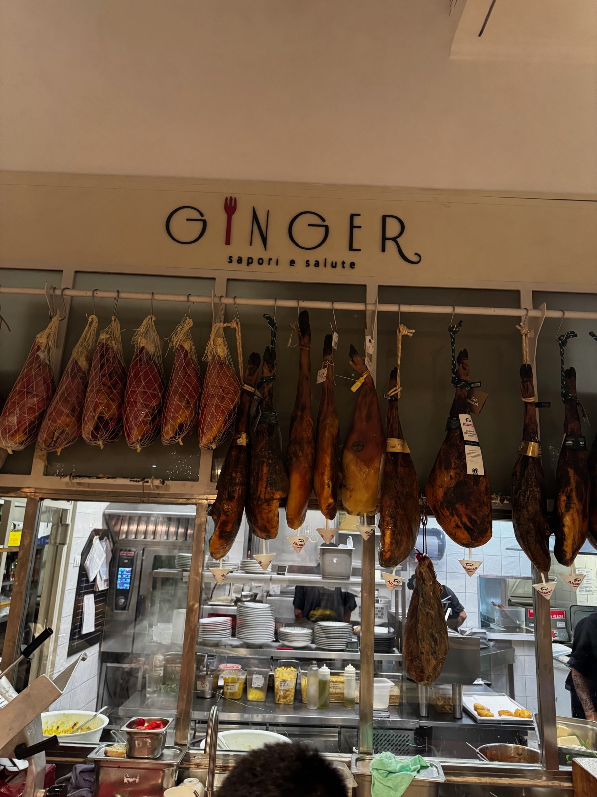 Ολόκληρα αλλαντικά ζαμπόν κρεμασμένα σε σειρές από μια μεταλλική σχάρα πάνω από έναν πάγκο ανοιχτής κουζίνας, μια ταμπέλα από πάνω που γράφει Ginger sapori e salute