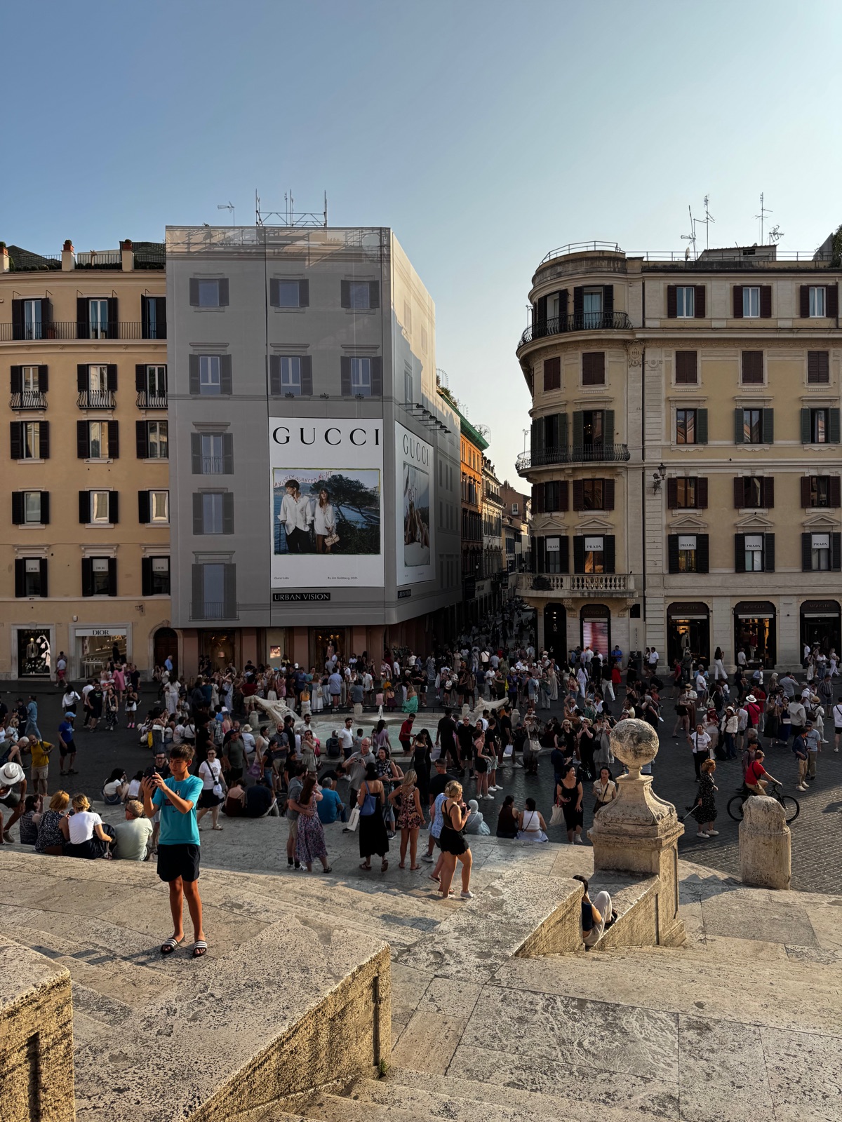 Η Piazza di Spagna από ψηλά το σούρουπο, εκατοντάδες άνθρωποι απλωμένοι στα σκαλιά και στην πλατεία, μια μεγάλη διαφημιστική πινακίδα στο απέναντι κτίριο