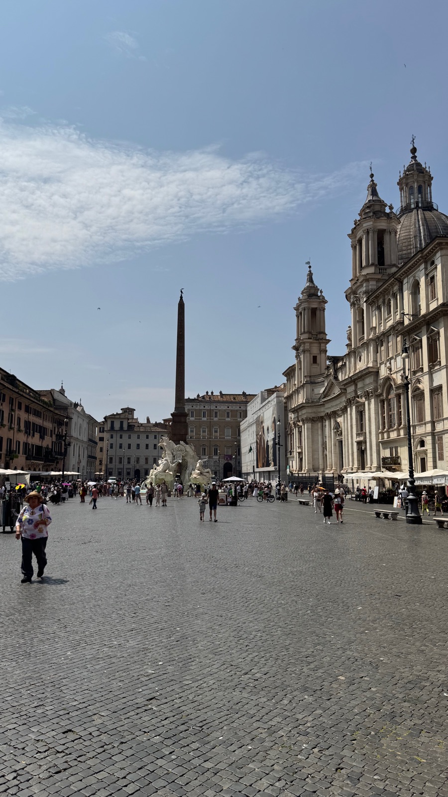 Η Piazza Navona ιδωμένη κατά μήκος της, ο οβελίσκος και το σιντριβάνι στη μία πλευρά, ώχρινα κτίρια και στις δύο πλευρές κάτω από έναν φωτεινό ουρανό