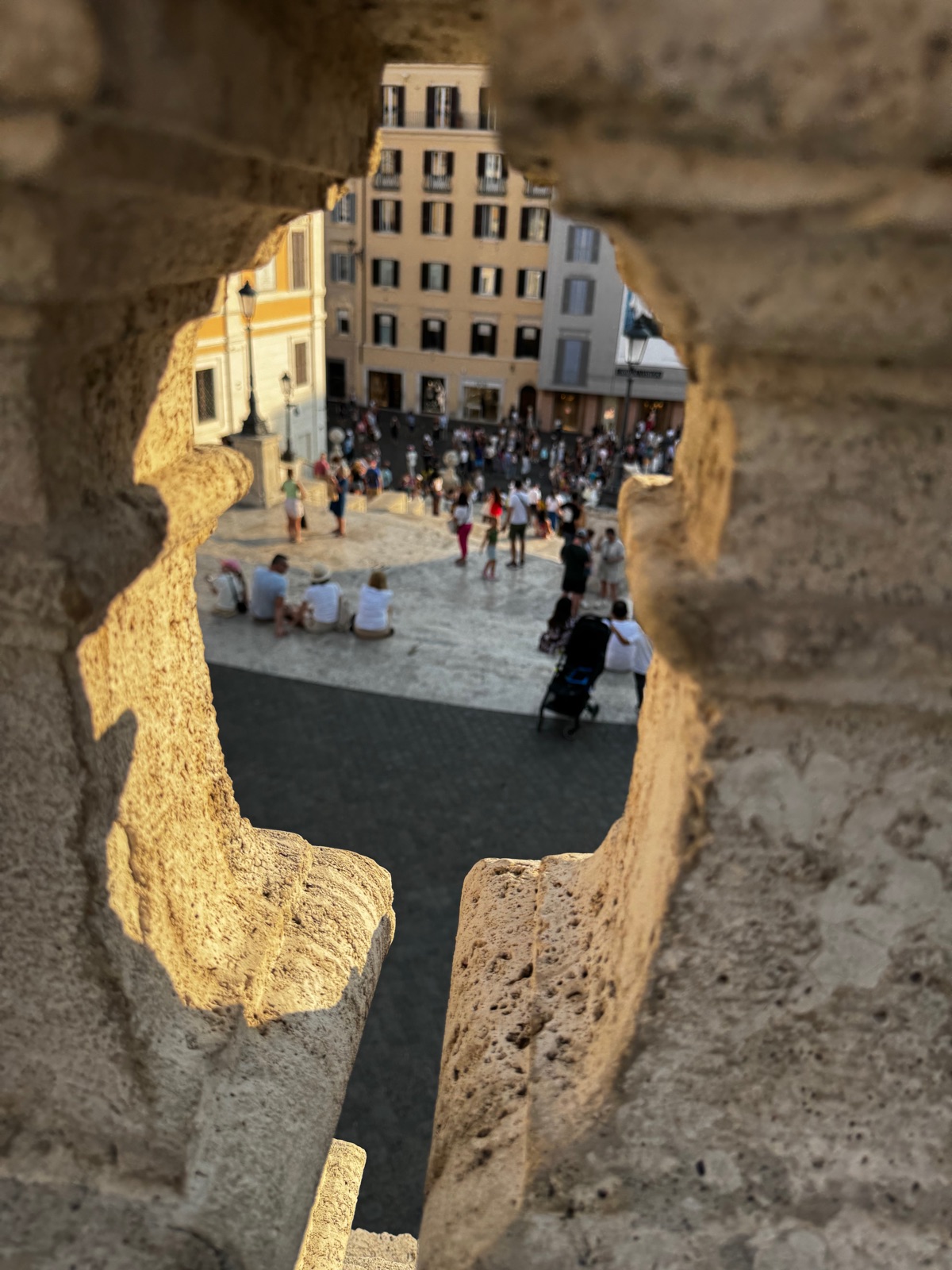 Η Piazza di Spagna ιδωμένη μέσα από ένα σπασμένο άνοιγμα σε φθαρμένο πέτρινο στηθαίο, το πλήθος και τα κτίρια καθαρά μέσα στο άνοιγμα, η πέτρα θολή γύρω