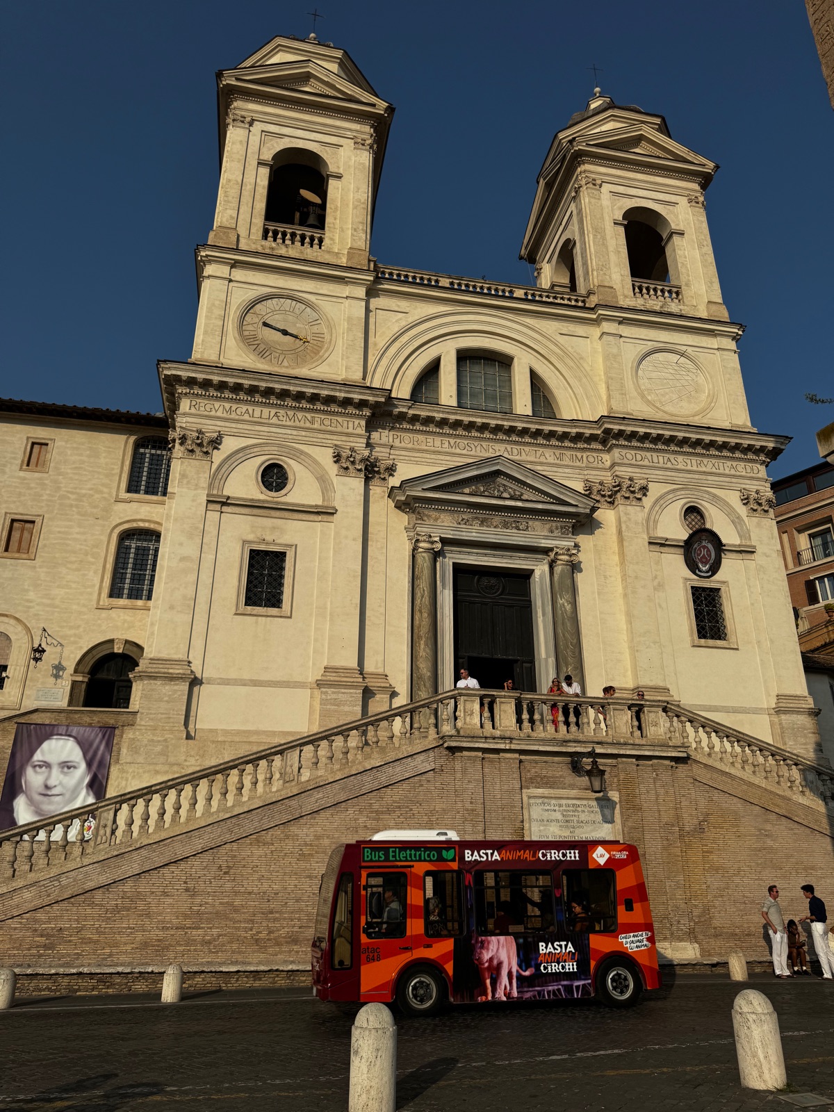 Η πρόσοψη της Trinita dei Monti με τους δίδυμους καμπαναριούς και το ρολόι της, ένα κόκκινο ανοιχτό τουριστικό λεωφορείο σταθμευμένο στον δρόμο από κάτω