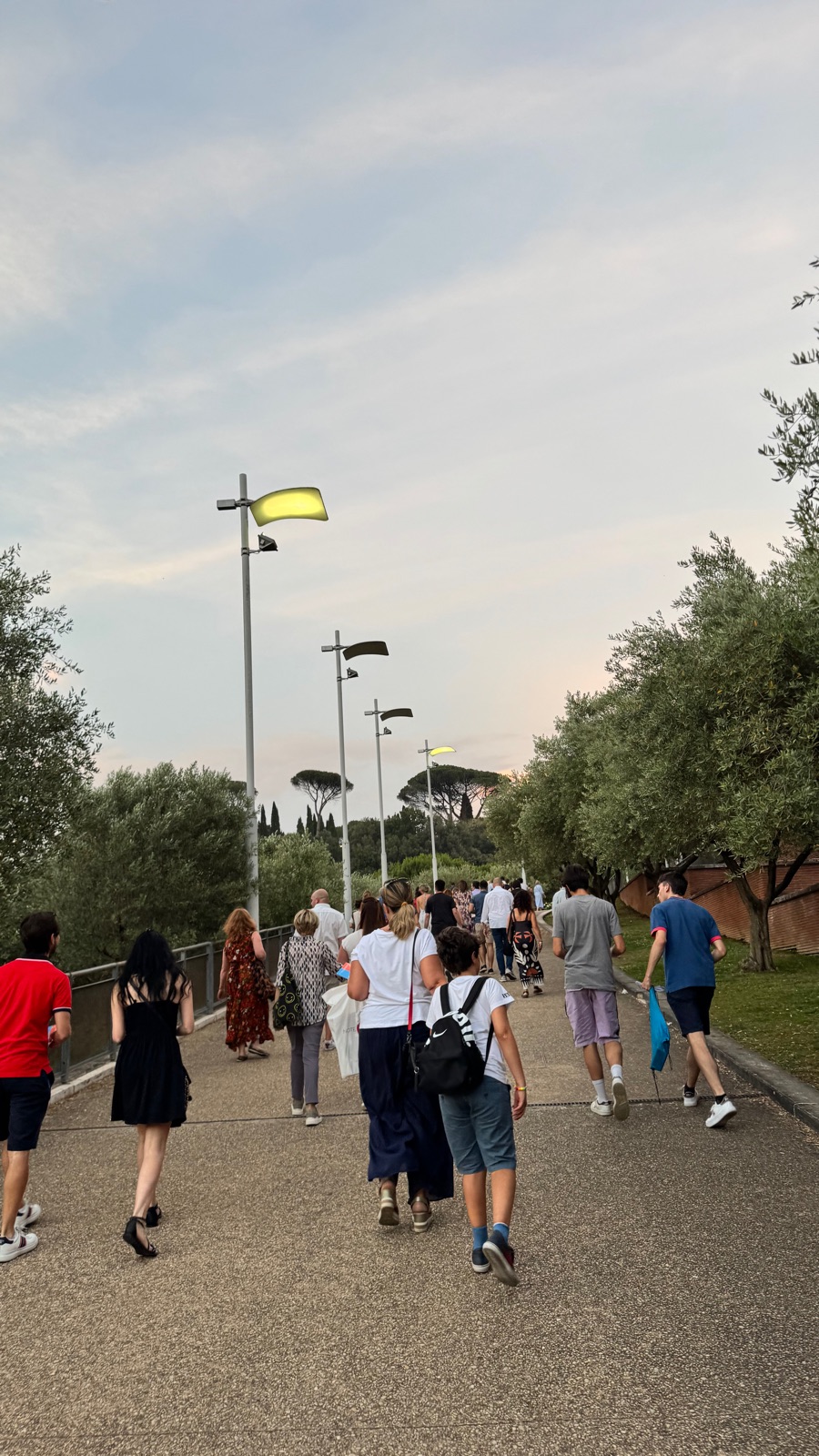 Άνθρωποι να περπατούν ένα δεντρόφυτο μονοπάτι το σούρουπο προς έναν χώρο συναυλιών, φανάρια αναμμένα πάνω σε έναν χλωμό βραδινό ουρανό