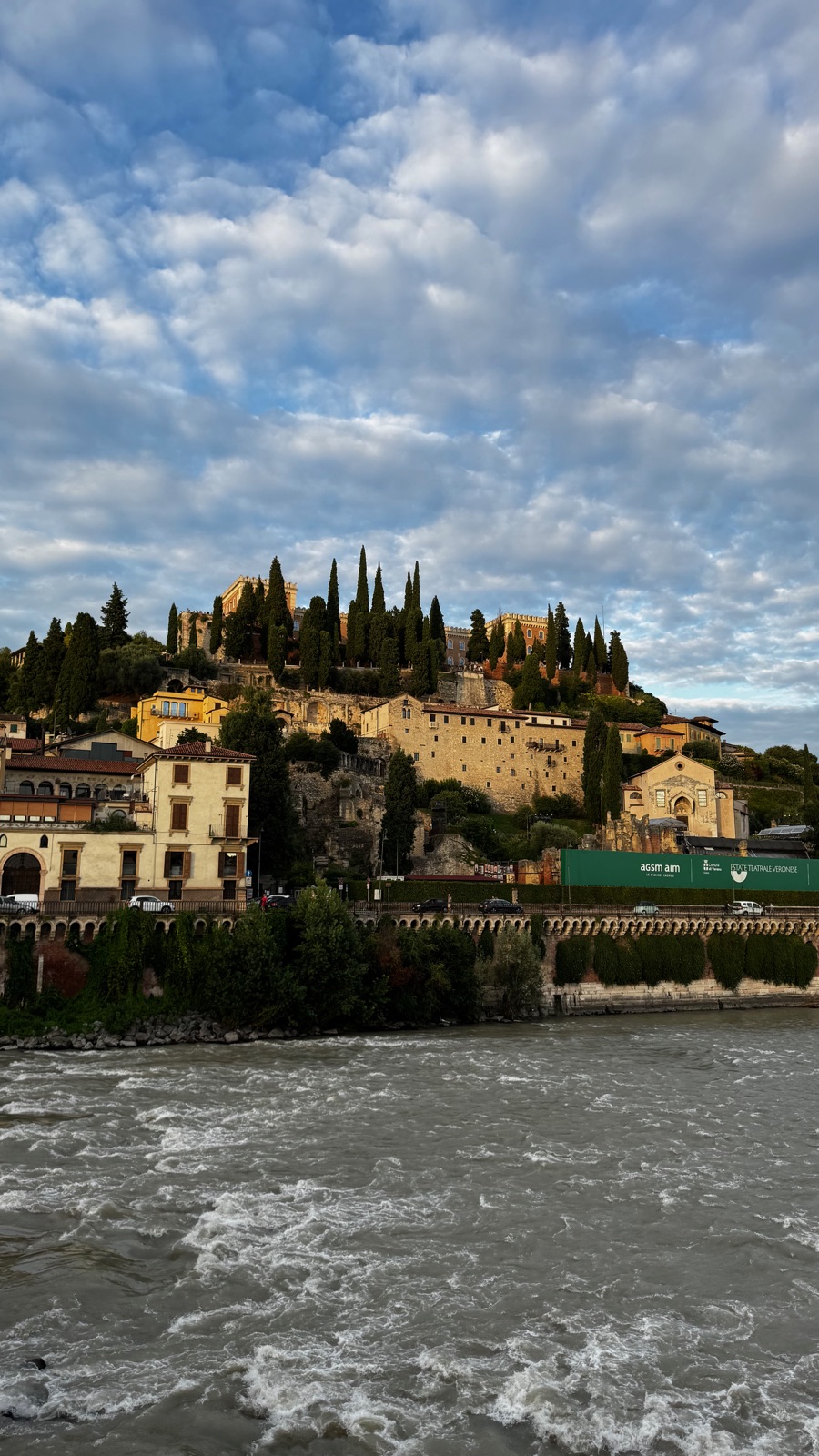 Ο λόφος του Castel San Pietro να λάμπει χρυσός σε χαμηλό βραδινό φως πάνω από τον ορμητικό Αδίγη, κυπαρίσσια στην κορυφή και μια πράσινη αφίσα θεατρικού φεστιβάλ στους πρόποδες