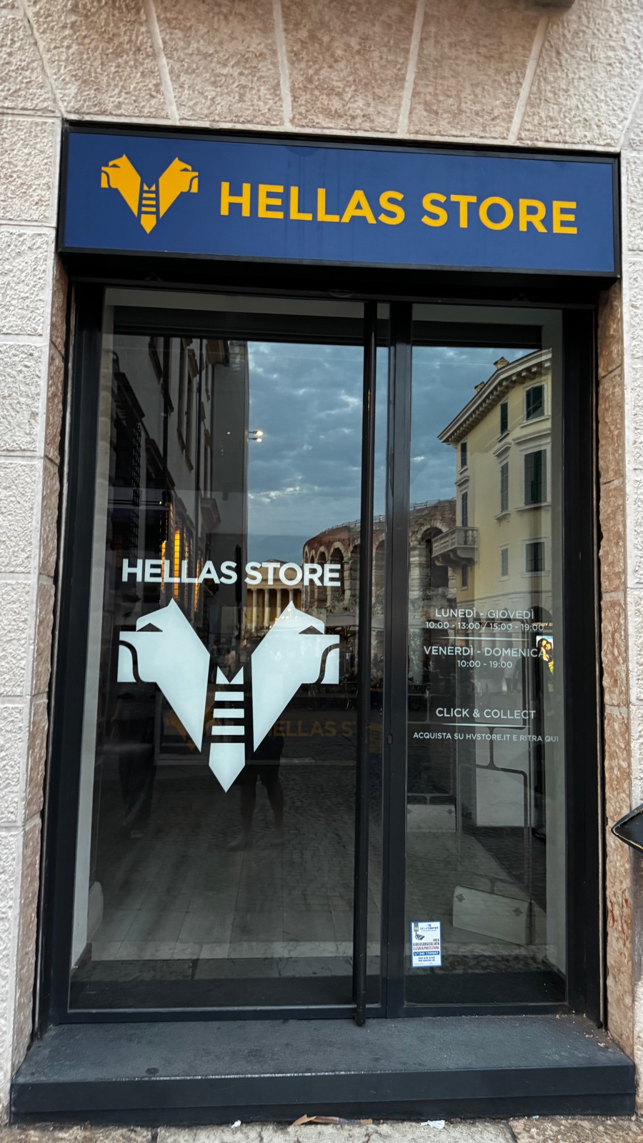 Η είσοδος του Hellas Store στη Βερόνα, μια σκούρα μπλε πινακίδα με το δικέφαλο σήμα του συλλόγου πάνω από την πόρτα και οι καμάρες της Αρένας καθρεφτισμένες στο τζάμι το σούρουπο