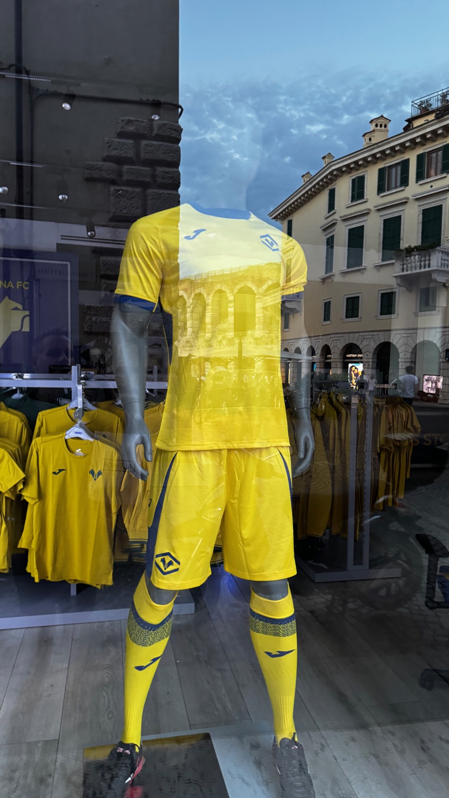 Μια κίτρινη εμφάνιση της Hellas Verona σε κούκλα στη βιτρίνα, ο ουρανός του σούρουπου και οι τοξοστοιχίες της Αρένας καθρεφτισμένες στο τζάμι πάνω από τη φανέλα