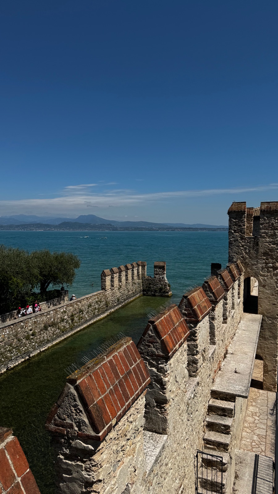 Ο περίδρομος κατά μήκος των επάλξεων της darsena με τούβλινες απολήξεις, η στεγασμένη κατασκευή της πύλης μπροστά και η λίμνη και η απέναντι όχθη πίσω